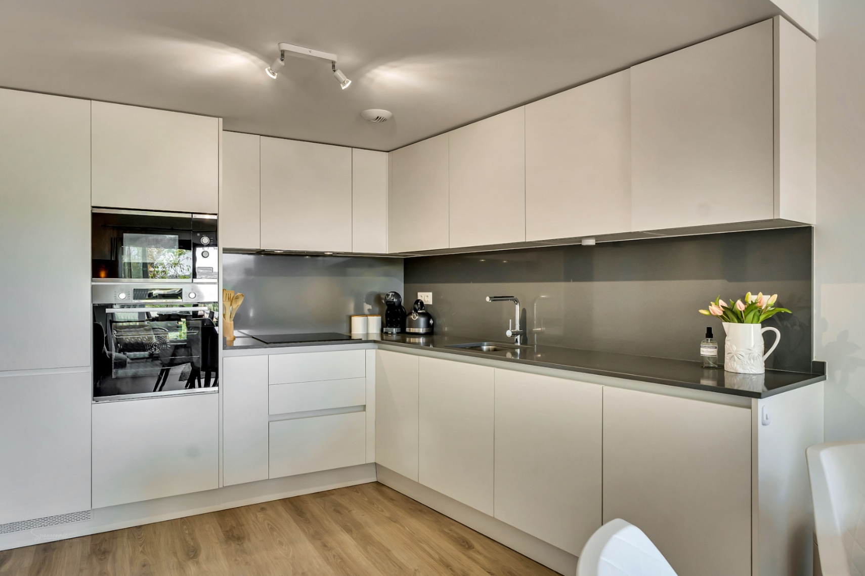 Cocina moderna en L con armarios blancos sin tiradores, encimera de cuarzo gris oscuro y electrodomésticos integrados. El suelo de madera aporta calidez al diseño minimalista.