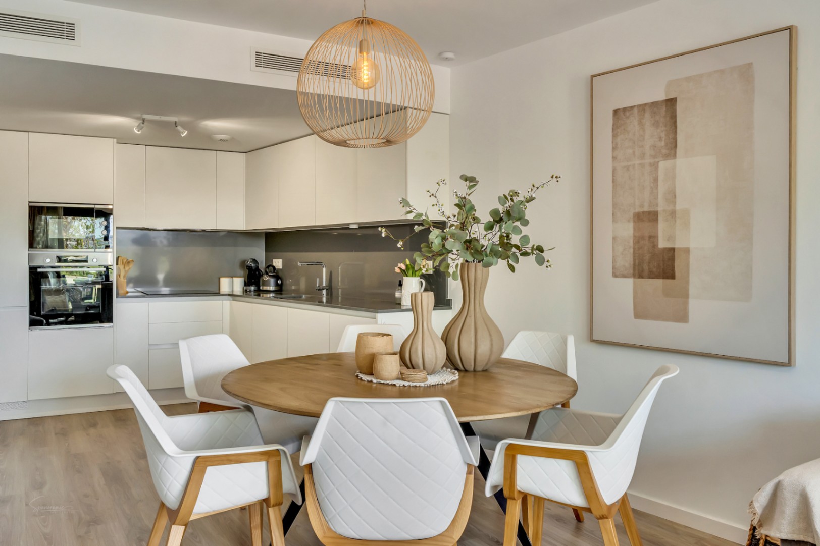 Comedor de concepto abierto con mesa circular de madera, sillas de diseño acolchadas y lámpara de mimbre, integrado con una cocina moderna de tonos neutros.