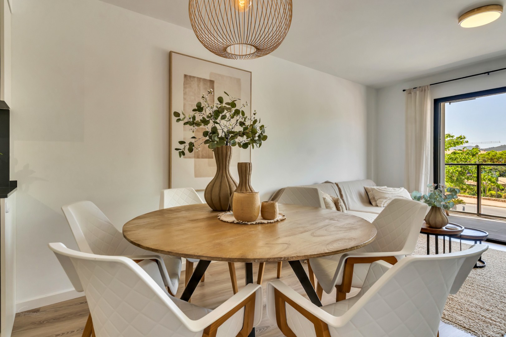 Comedor de planta abierta con mesa circular de madera, sillas de diseño acolchadas y una lámpara de mimbre, integrado en un salón luminoso con acceso directo al balcón.