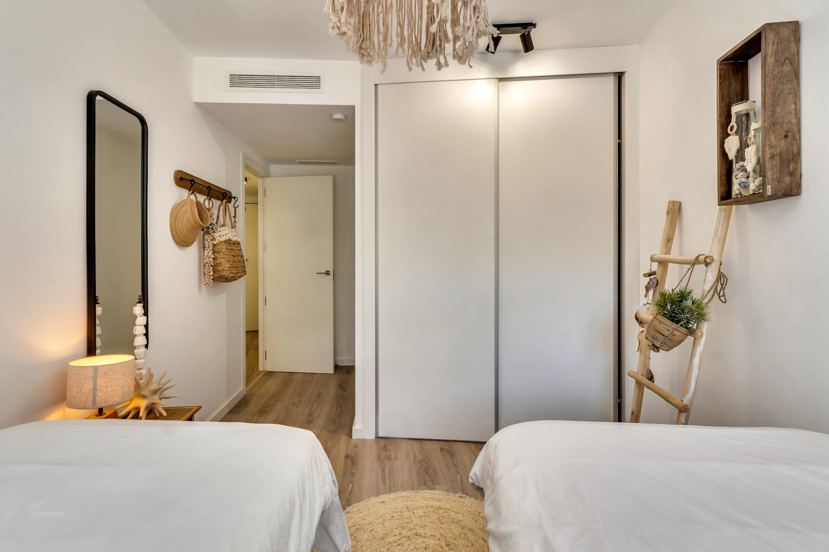 Dormitorio con dos camas individuales, armario empotrado de puertas correderas blancas y suelos de madera, decorado con sutiles toques mediterráneos y bohemios.