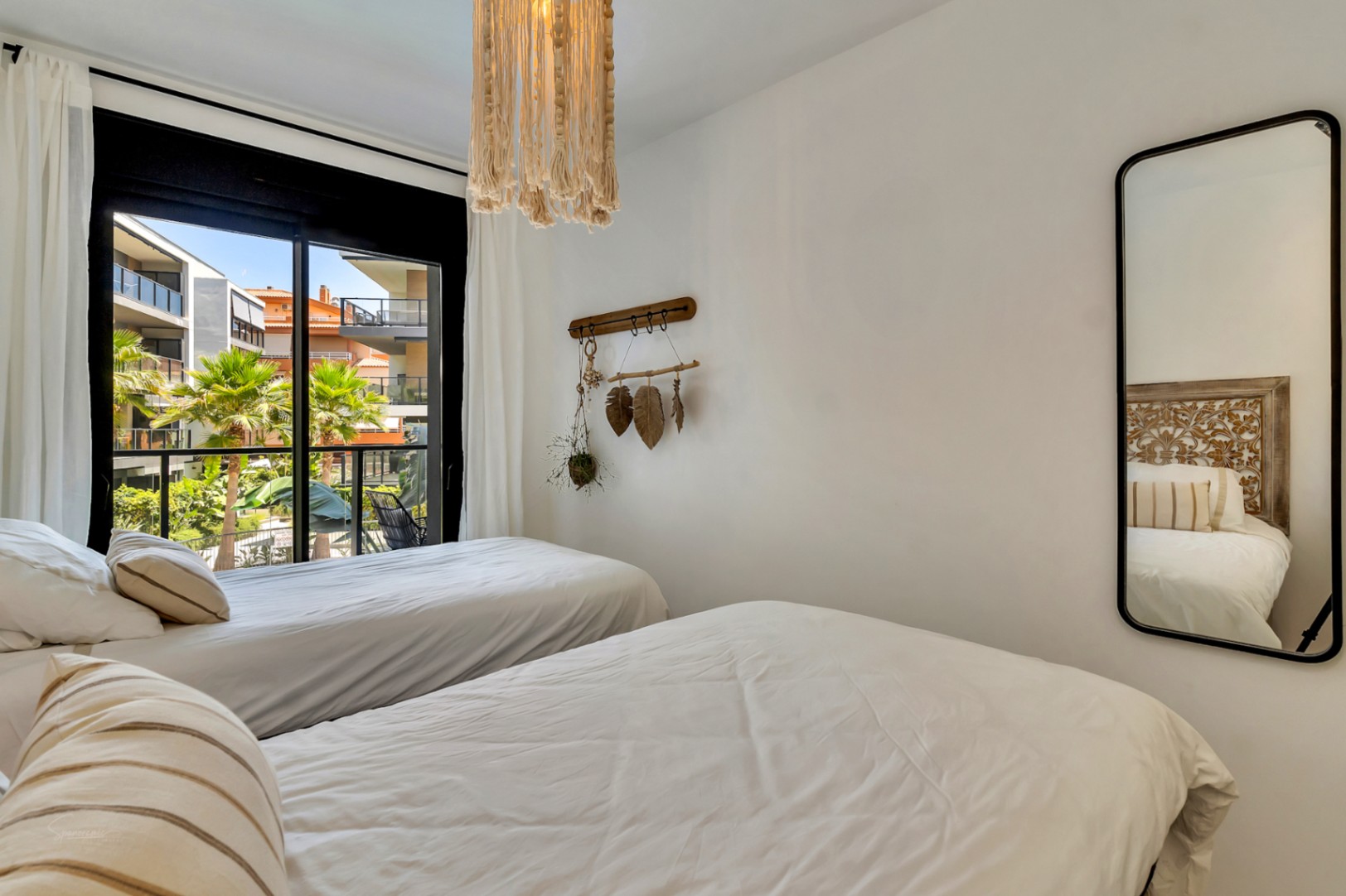 Dormitorio con dos camas individuales, lámpara de macramé y espejo vertical. El ventanal muestra vistas despejadas a la urbanización y zonas verdes de la propiedad.