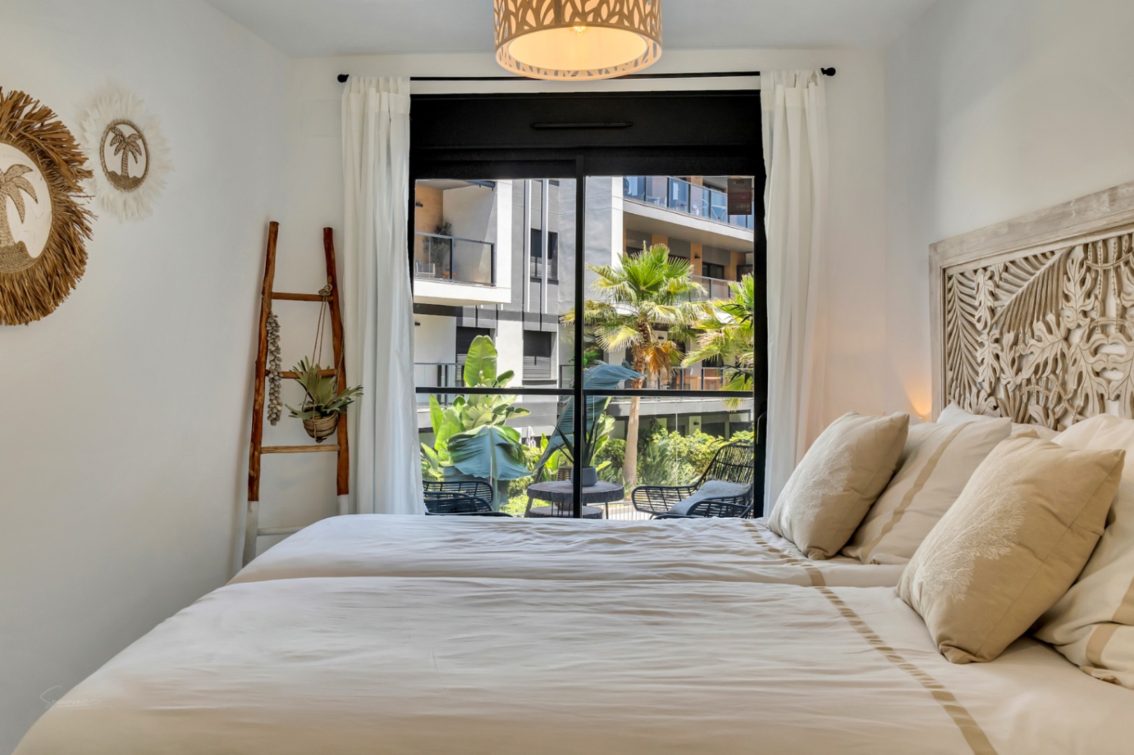 Dormitorio luminoso con cabecero de madera tallada, decoración de estilo mediterráneo y acceso directo a una terraza privada con vistas a las zonas comunes.