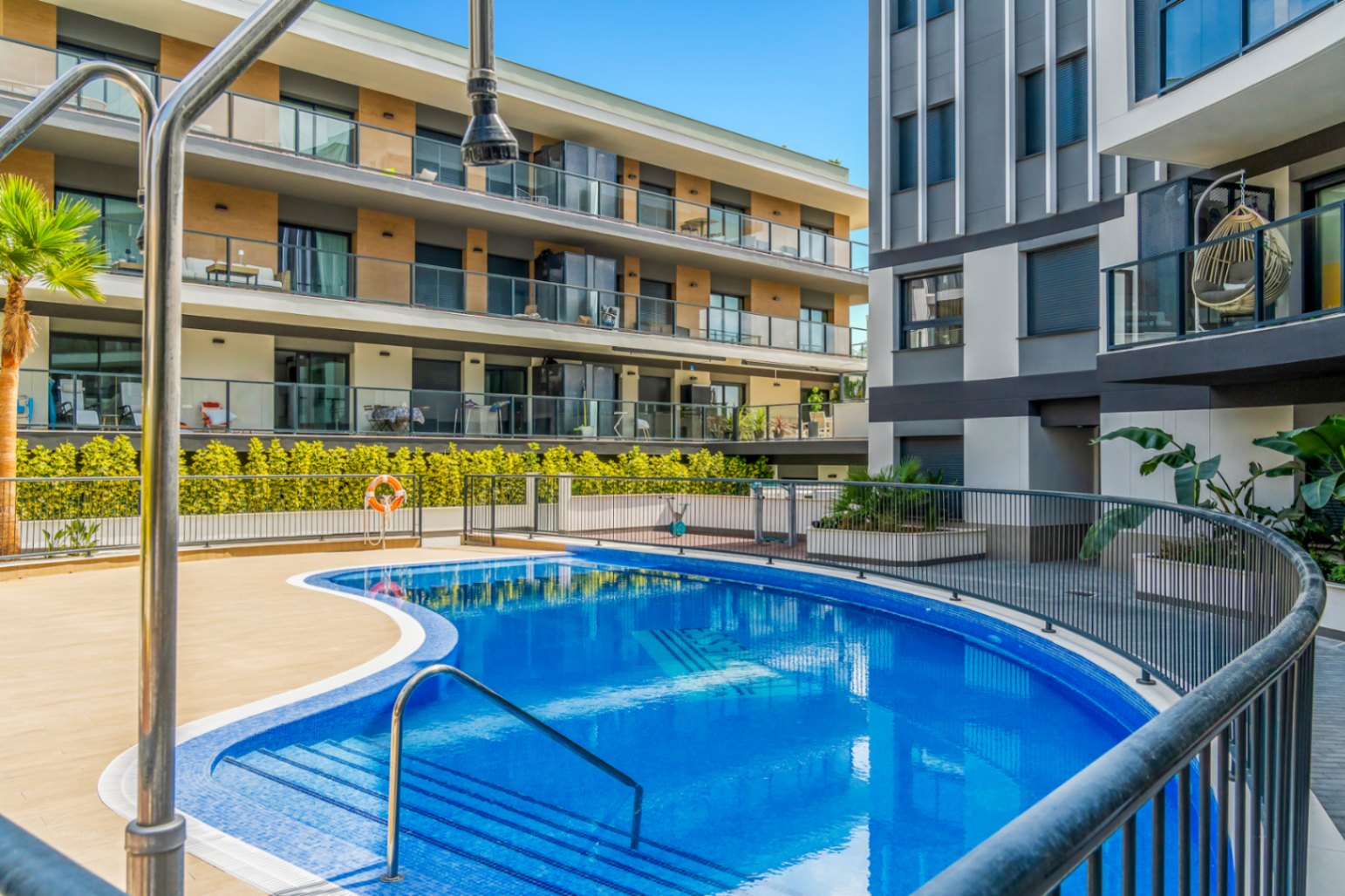 Piscina comunitaria de diseño curvo rodeada de una terraza solárium y vallado de seguridad, situada frente a la fachada contemporánea del complejo residencial en Jávea.