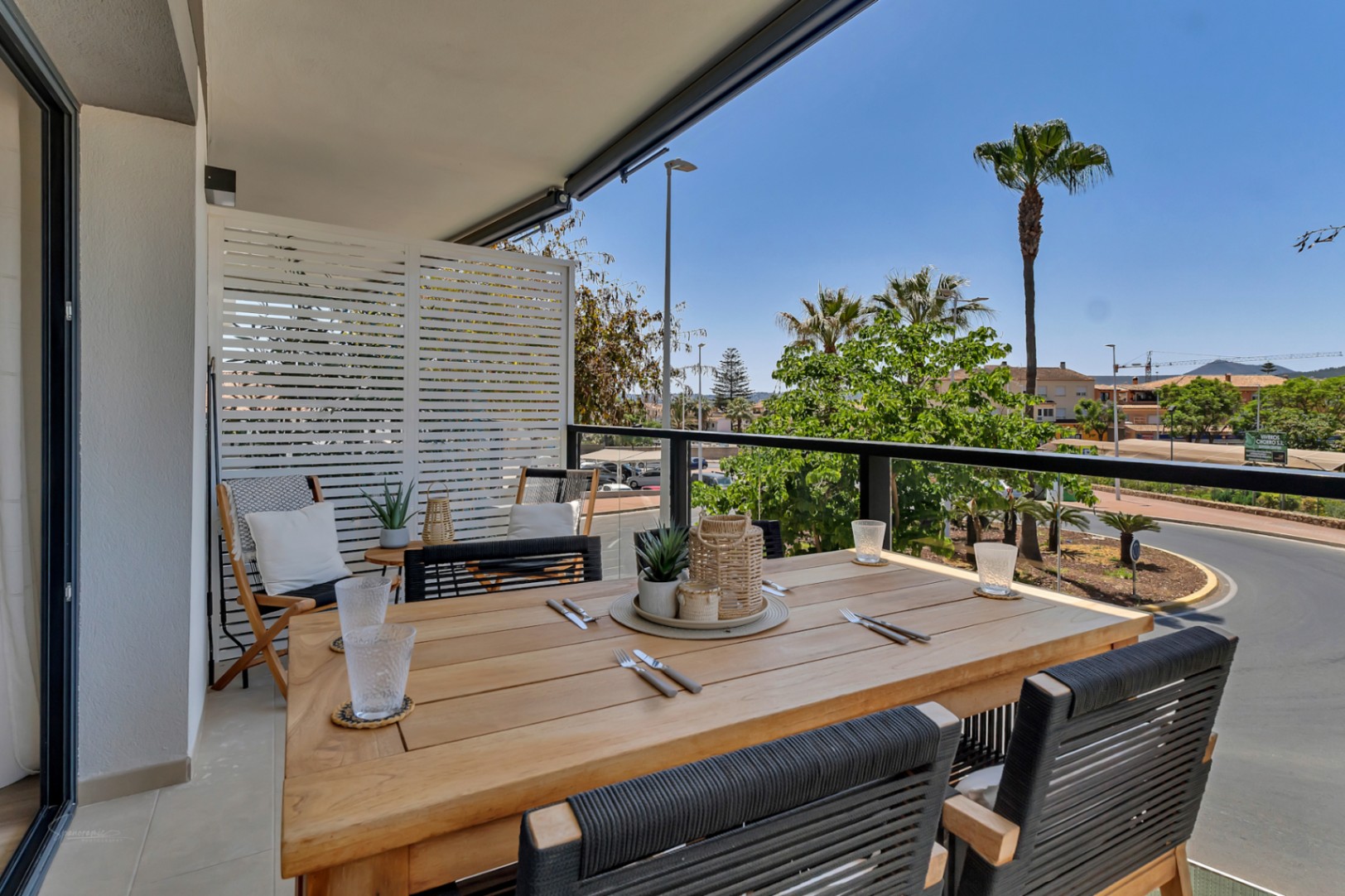 Terraza cubierta con mesa de comedor de madera, sillas de cuerda negra y vistas despejadas al entorno urbano y las palmeras de Jávea.