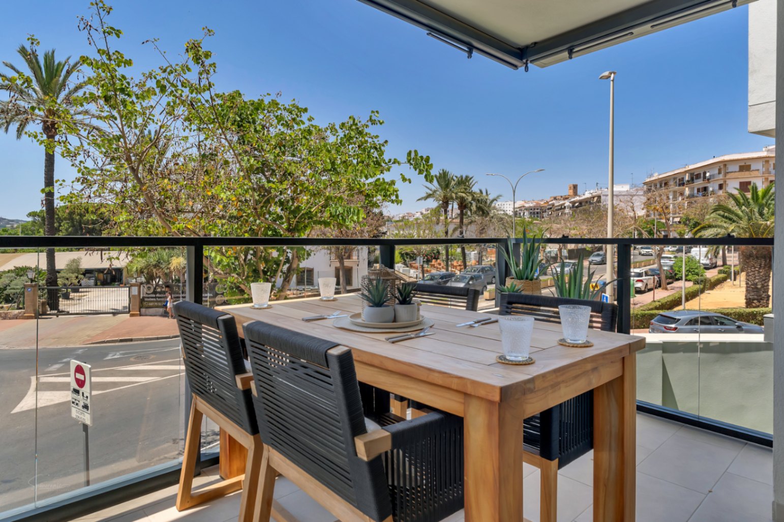 Terraza privada con mesa de comedor de madera y sillas de cuerda trenzada, protegida por una barandilla de cristal con vistas despejadas al entorno urbano.