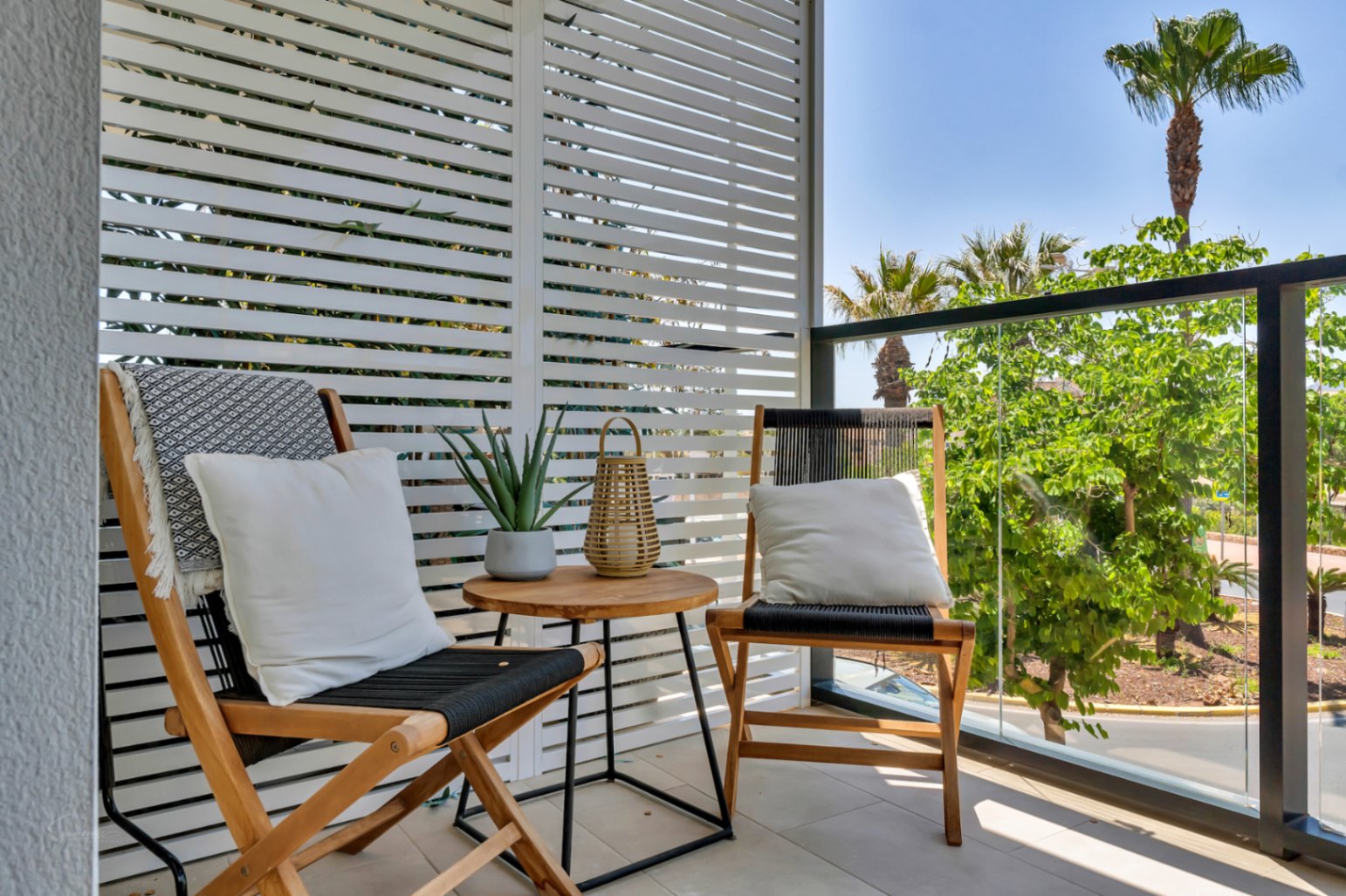 Terraza privada con suelo cerámico, mobiliario de madera y cuerda, protegida por celosía blanca y barandilla de cristal con vistas a zonas verdes.