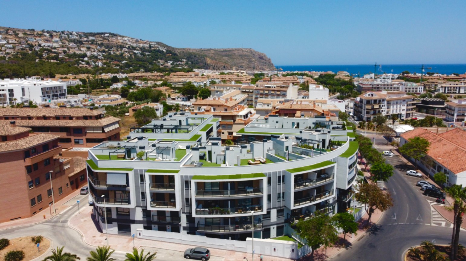 Vista aérea del complejo residencial contemporáneo en Jávea, destacando su arquitectura curva, terrazas con césped artificial y vistas panorámicas al mar Mediterráneo.