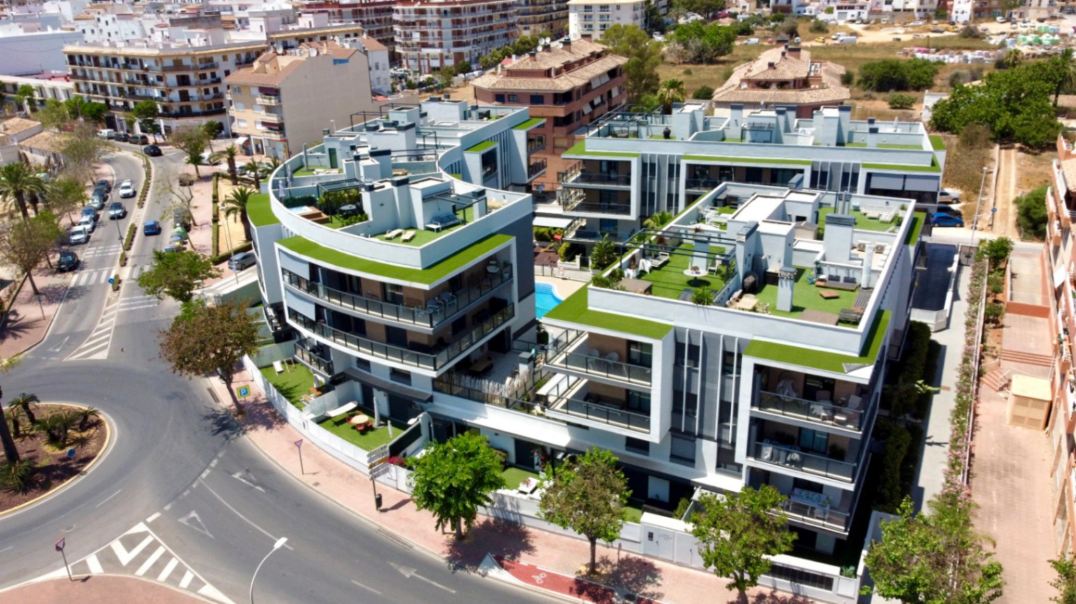 Vista aérea del complejo residencial moderno con arquitectura curva, azoteas ajardinadas y piscina central, ubicado en una zona urbana estratégica de Jávea.