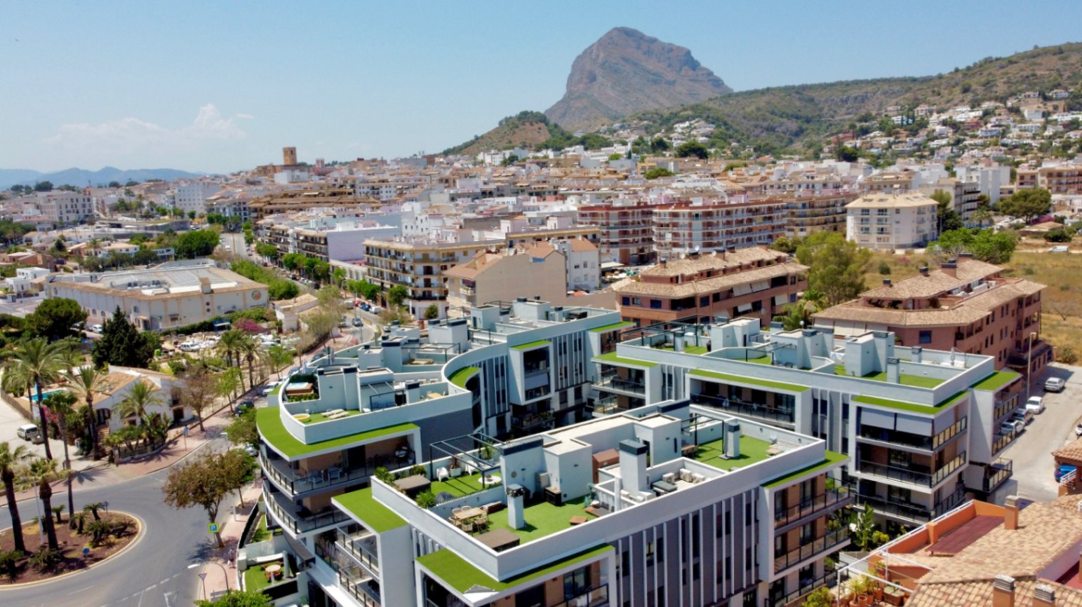 Vista aérea del complejo residencial moderno en Jávea, destacando sus azoteas con césped artificial y la emblemática montaña del Montgó al fondo bajo un cielo despejado.
