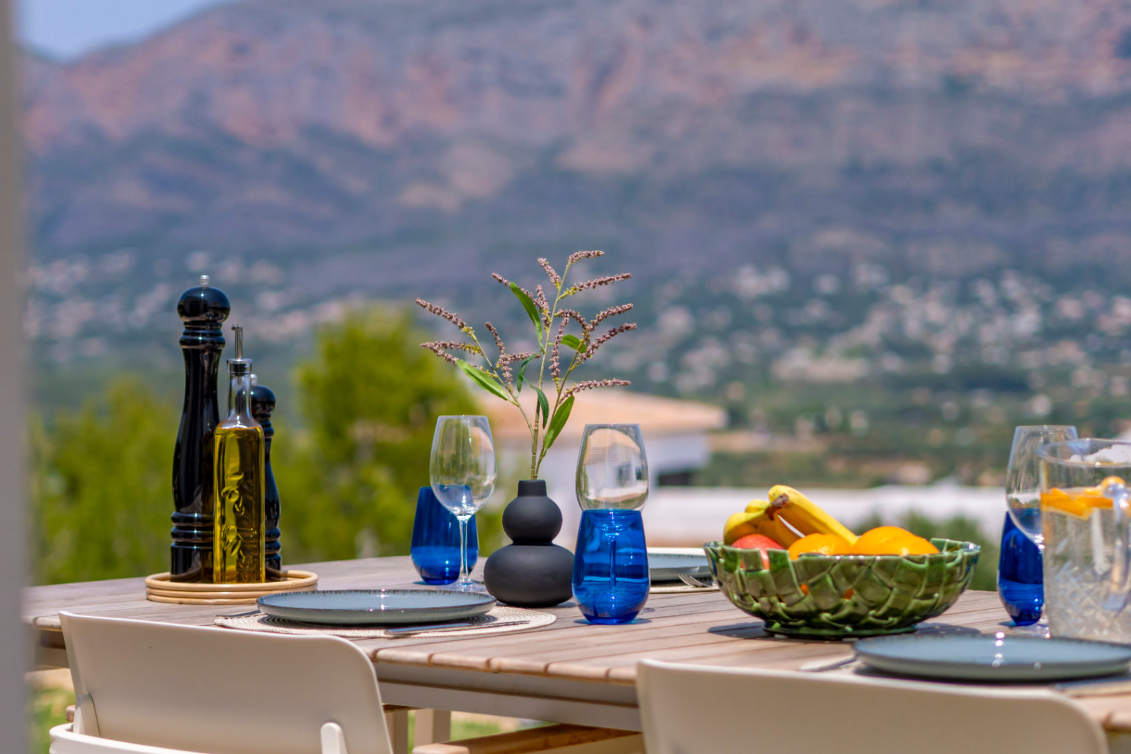 Comedor al aire libre con vistas a la montaña. Mesa de madera con vajilla azul y frutero. Ambiente mediterráneo.