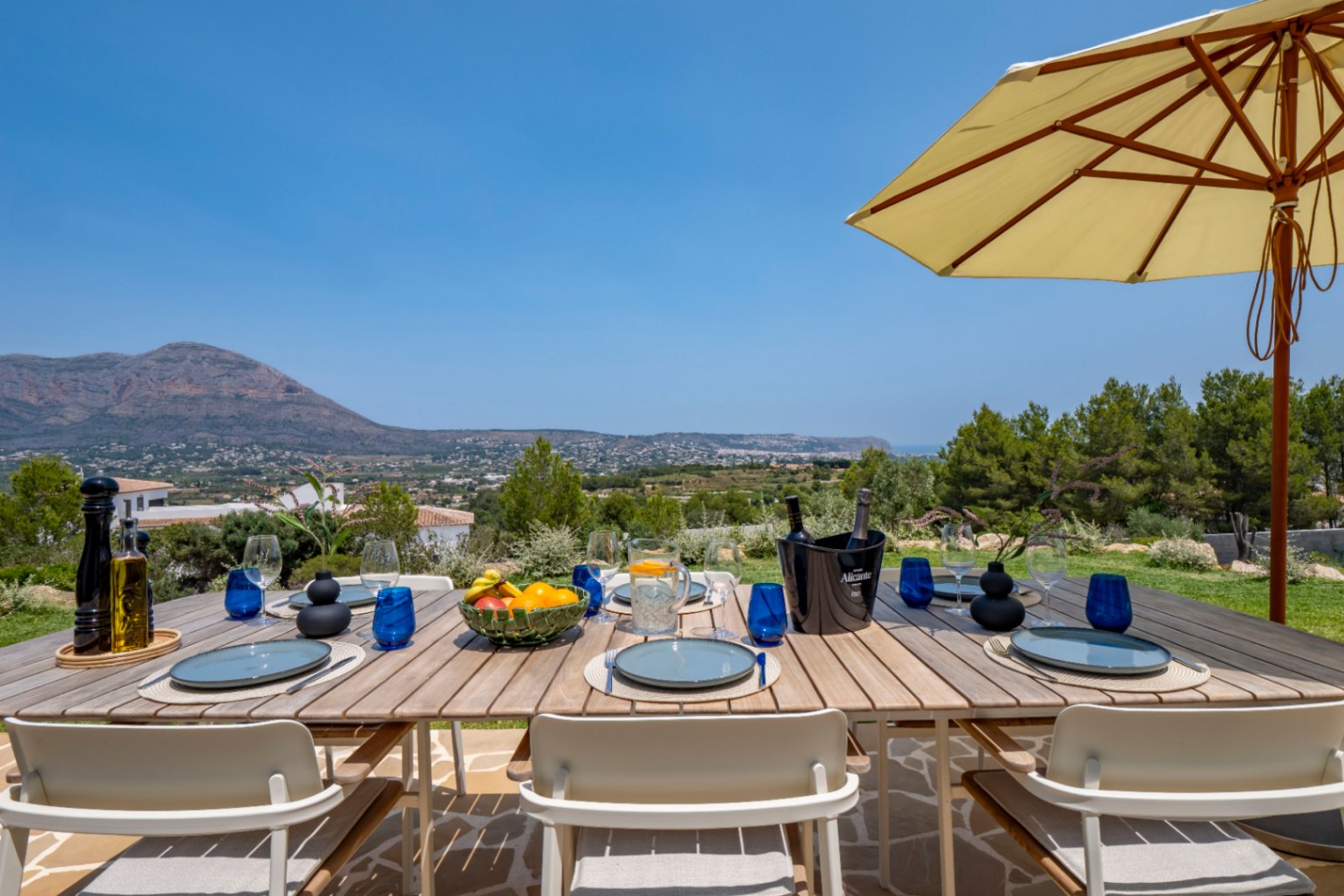Comedor exterior con mesa de madera, vajilla azul y vistas panorámicas a la montaña en Jávea.