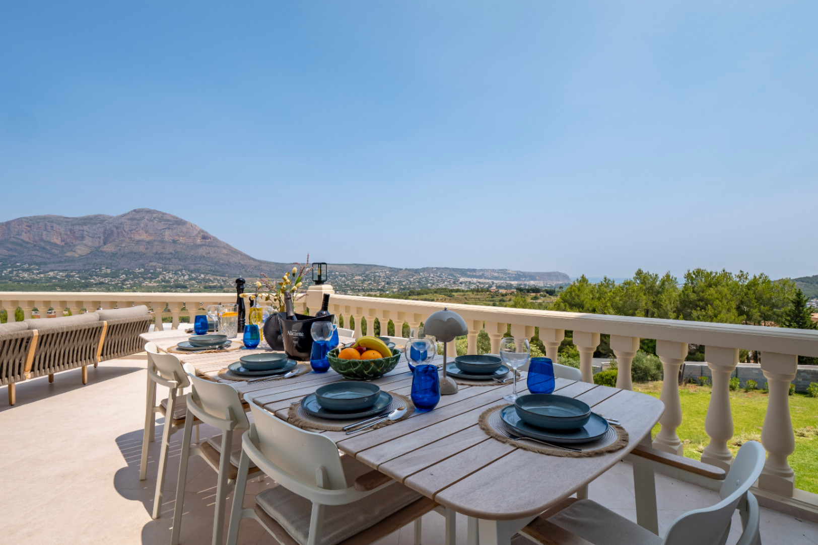 Comedor exterior con mesa de madera y vistas panorámicas a la montaña y la costa desde la terraza.