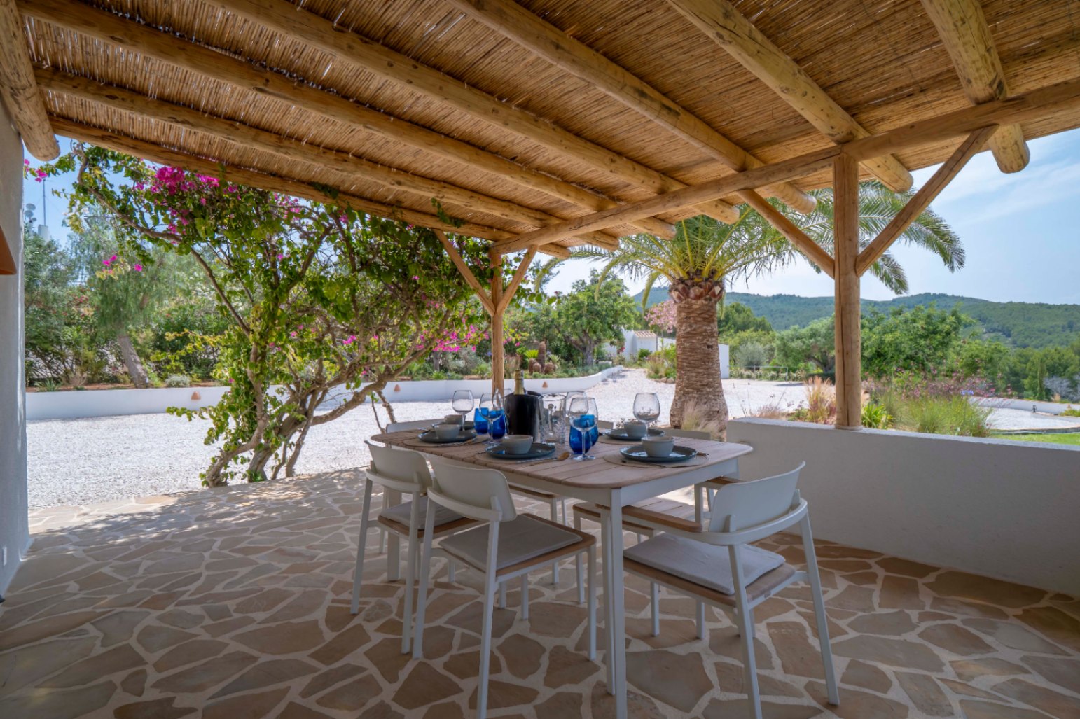 Comedor exterior cubierto con techo de paja, suelo de piedra irregular y vistas al jardín mediterráneo.
