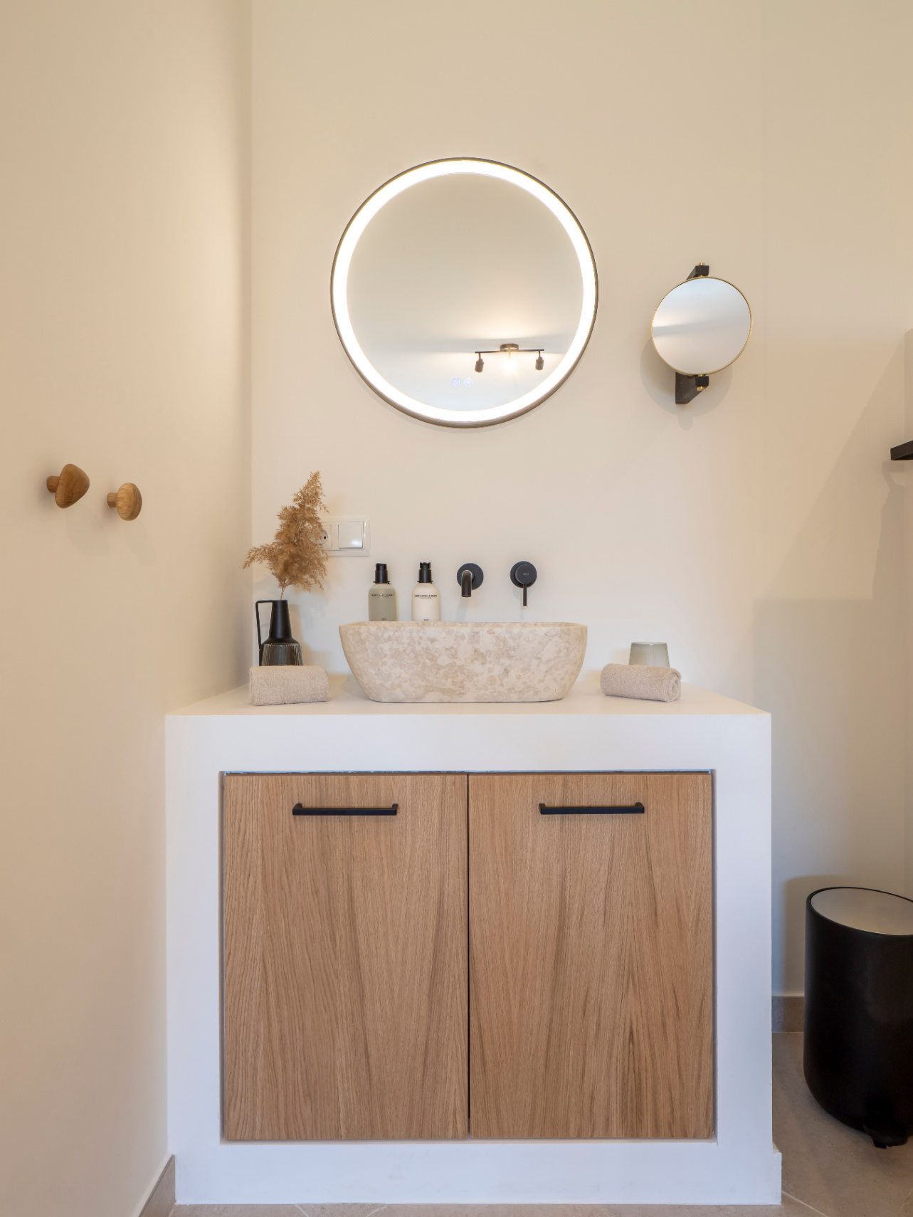Detalle de baño con lavabo de piedra natural, espejo circular retroiluminado y mueble de madera clara con tiradores negros.