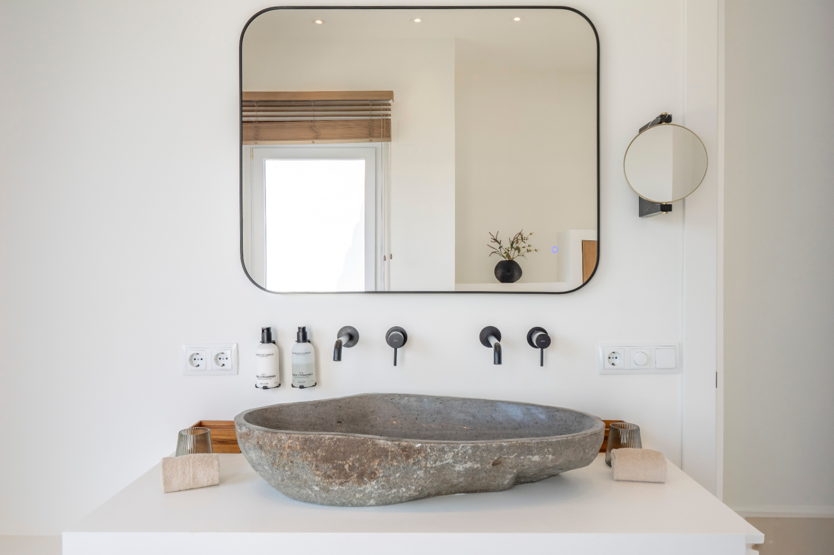 Detalle de baño con lavabo de piedra natural y grifería negra mate. Espejo rectangular con luz integrada.