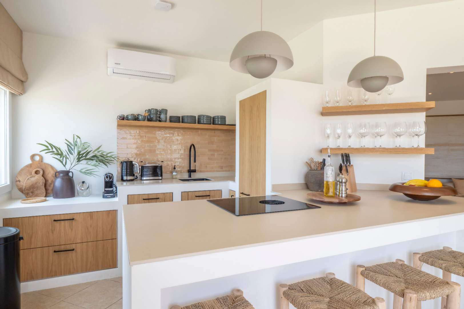 Detalle de la cocina con isla central, taburetes de madera y encimera clara. Estantes con cristalería y menaje.