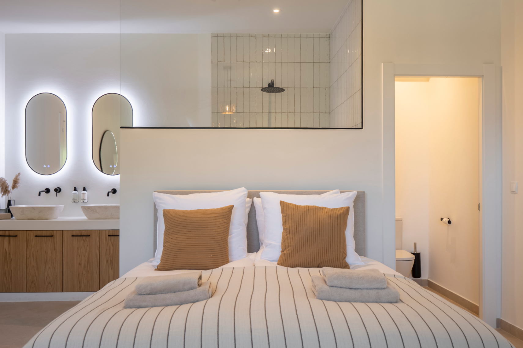 Dormitorio con baño en suite. Lavabos de piedra sobre mueble de madera clara e iluminación LED.