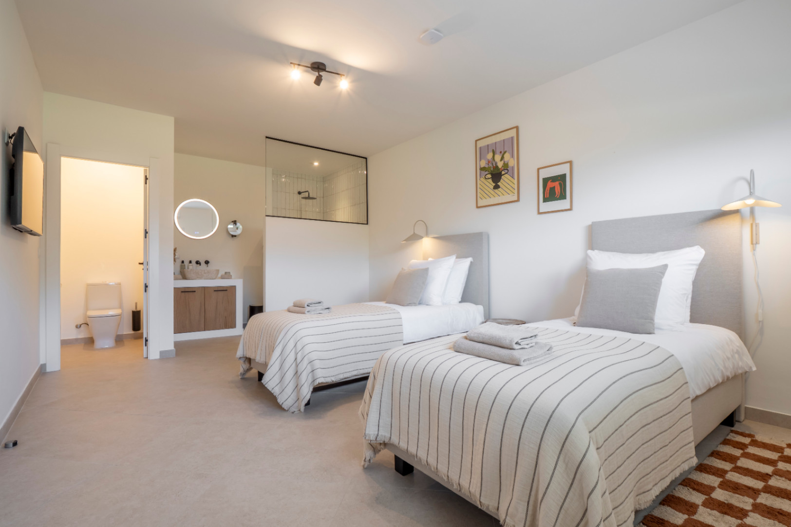 Dormitorio luminoso con dos camas individuales, baño en suite con ducha acristalada y tocador de madera clara.