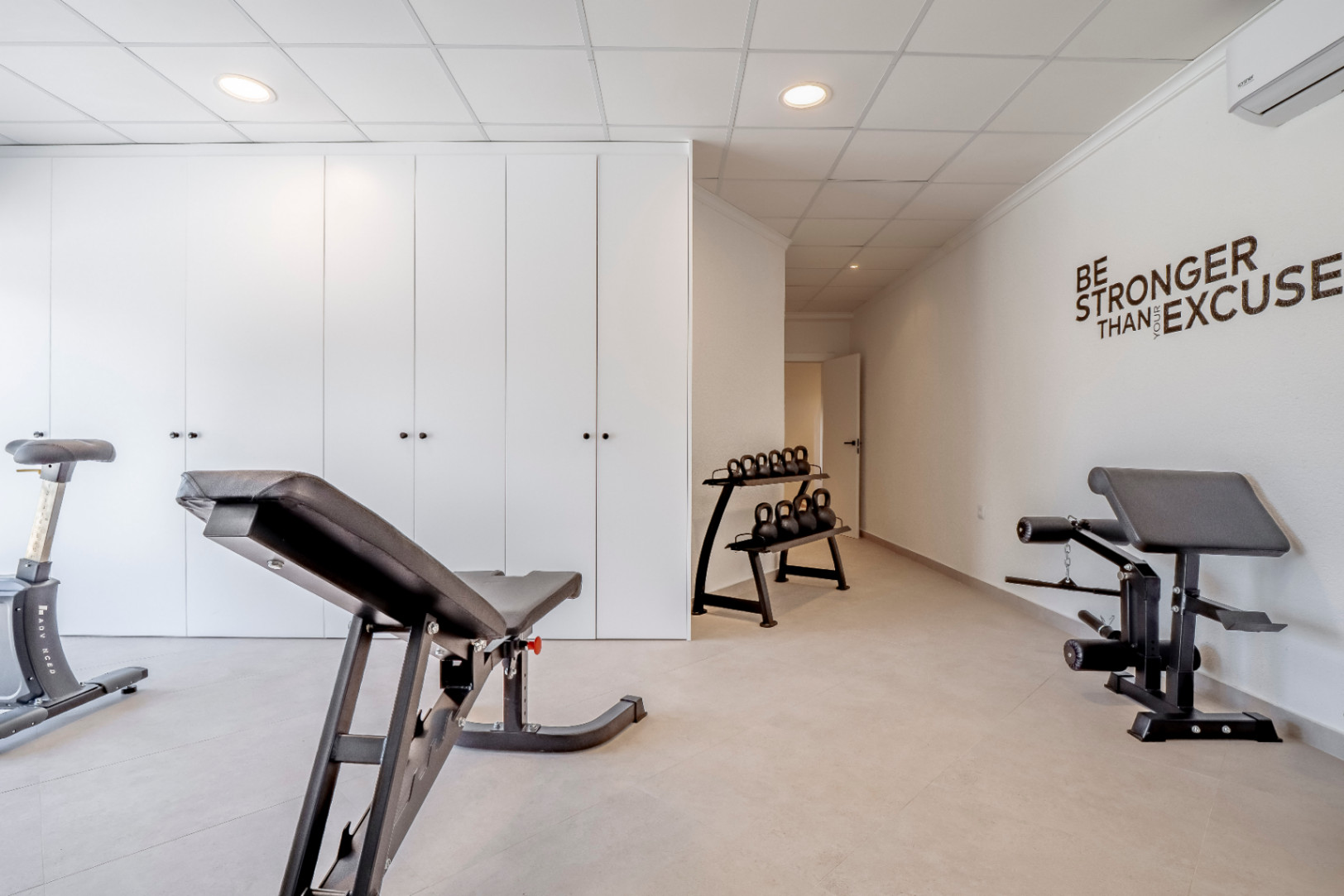 Gimnasio con suelo cerámico, paredes blancas y armarios empotrados. Equipo de entrenamiento con pesas y banco.