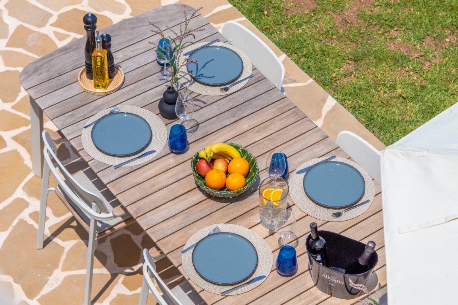 Mesa de comedor exterior con platos azules, cristalería y frutero, lista para disfrutar en el jardín.