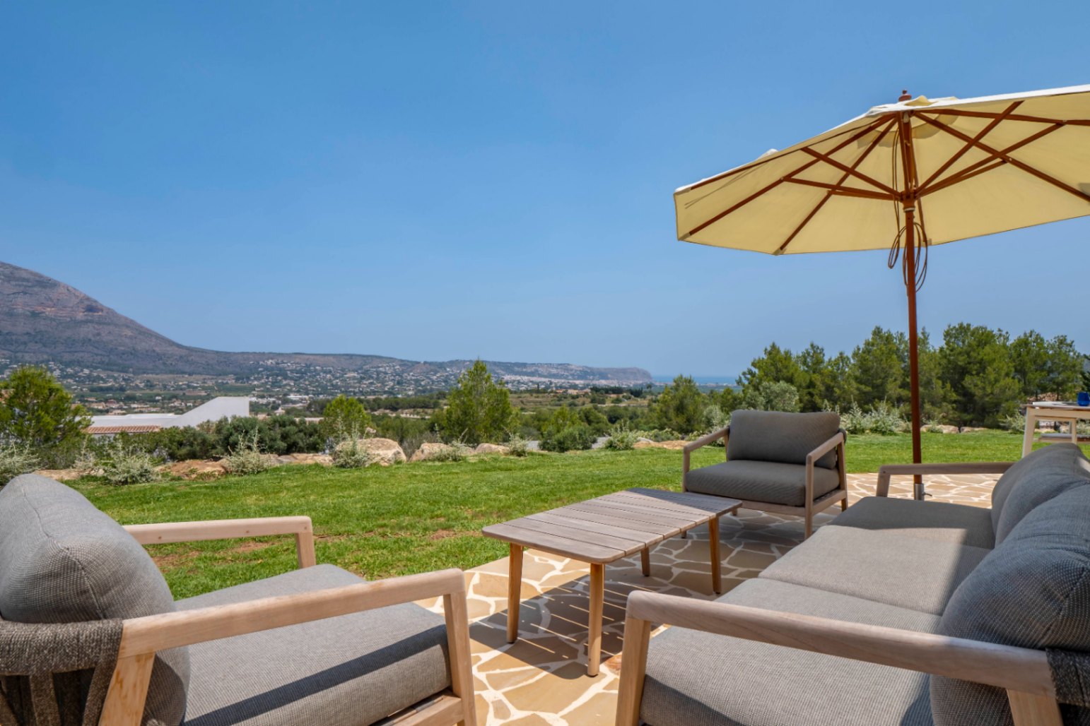 Terraza con vistas: mobiliario de exterior en piedra y madera bajo sombrilla, con paisaje de Jávea.