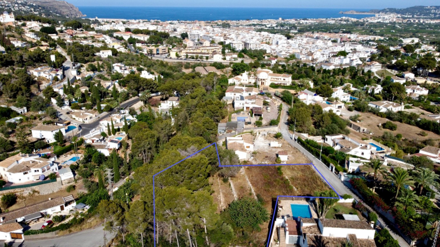 Vista aérea de la parcela de 1.908 m² delimitada en azul, con pinada propia y vistas panorámicas al mar y al casco urbano de Jávea.