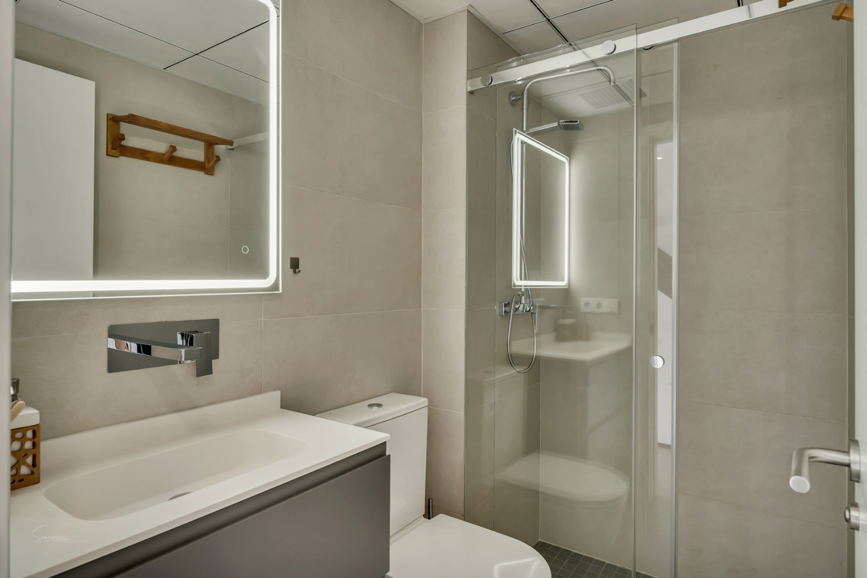 Baño moderno con azulejos neutros, mueble de lavabo gris y espejo con iluminación LED perimetral junto a una ducha con mampara de cristal corredera.