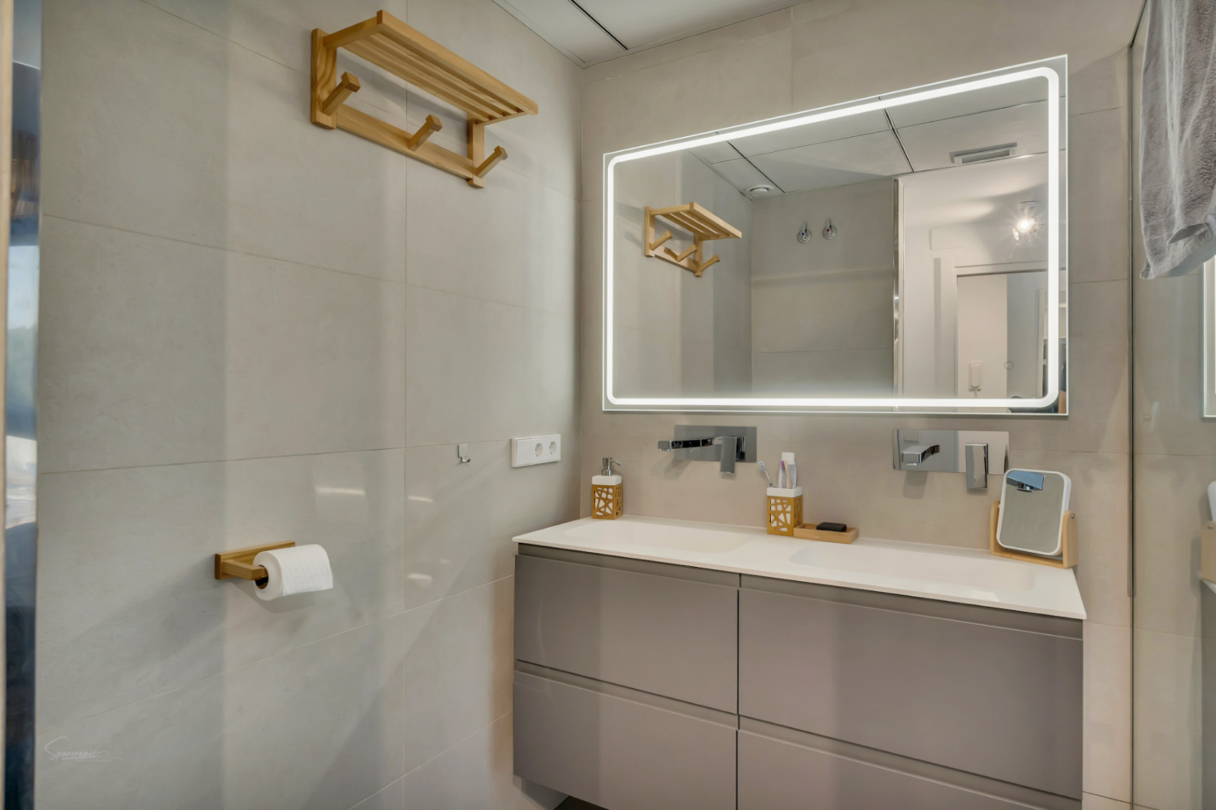 Baño moderno con lavabo de doble seno, mueble gris suspendido y espejo con iluminación LED perimetral. Destacan los accesorios de madera y el revestimiento en tonos neutros.