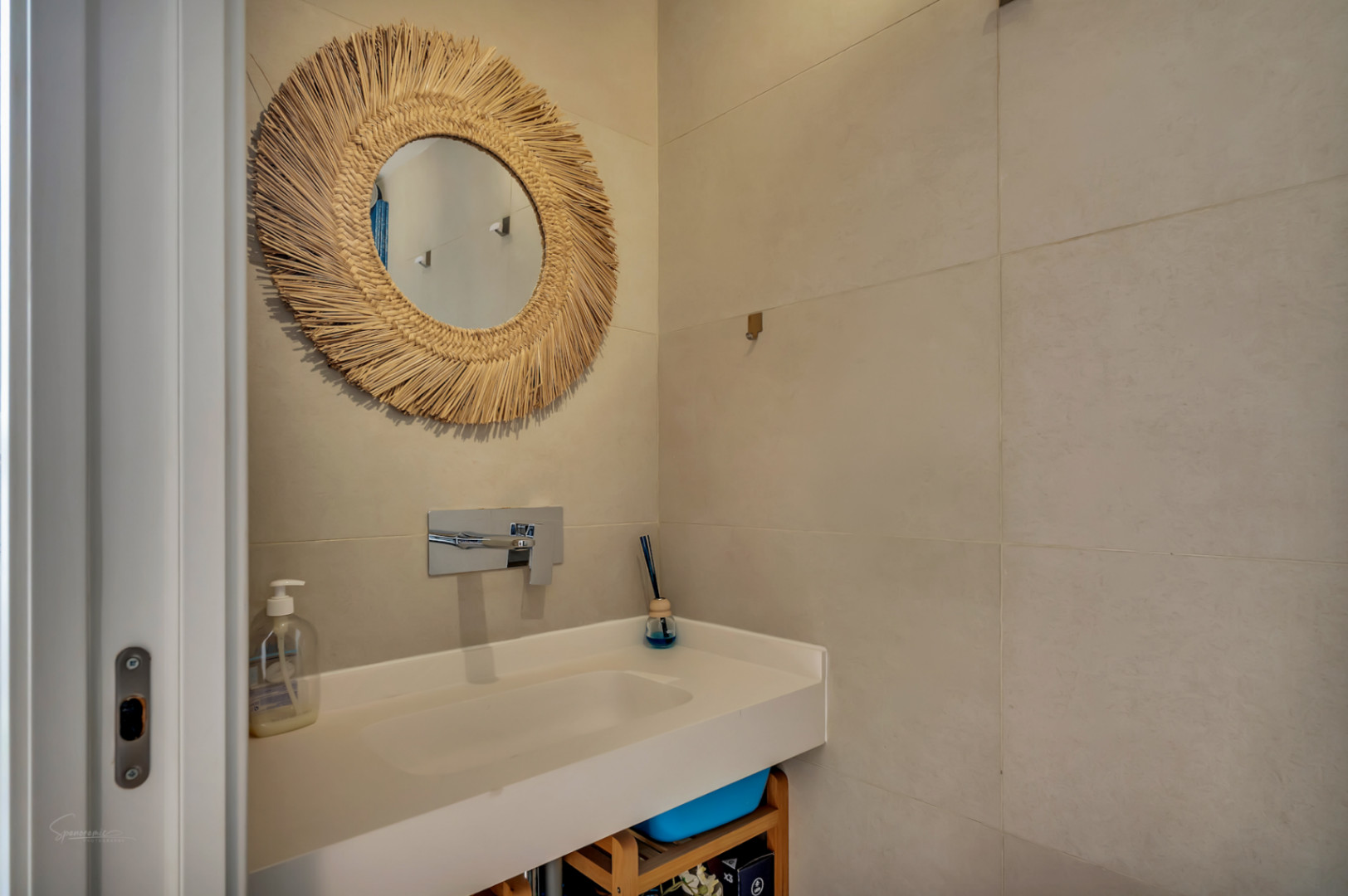 Baño moderno con lavabo integrado blanco, grifería cromada empotrada y un espejo decorativo con marco de fibras naturales sobre azulejos de gran formato en tonos arena.