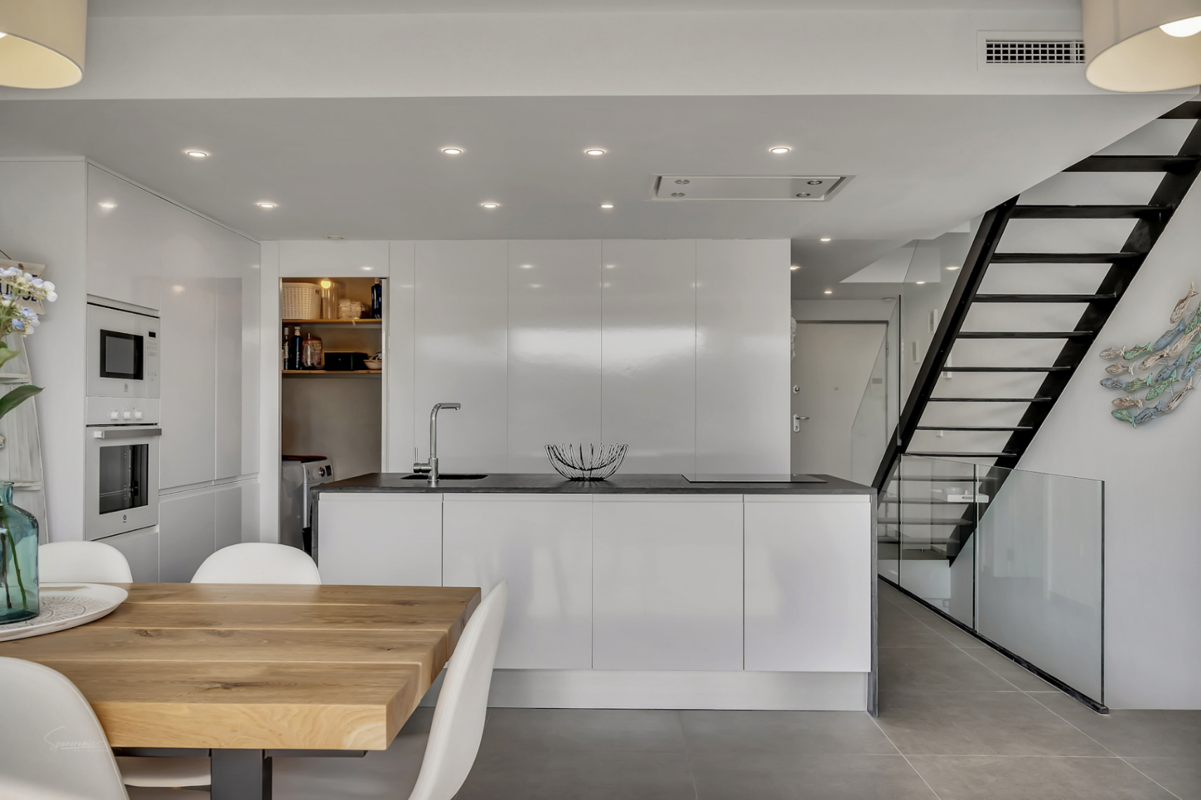 Cocina moderna de concepto abierto con isla central, armarios blancos de alto brillo y electrodomésticos integrados junto a una escalera de diseño industrial y cristal.