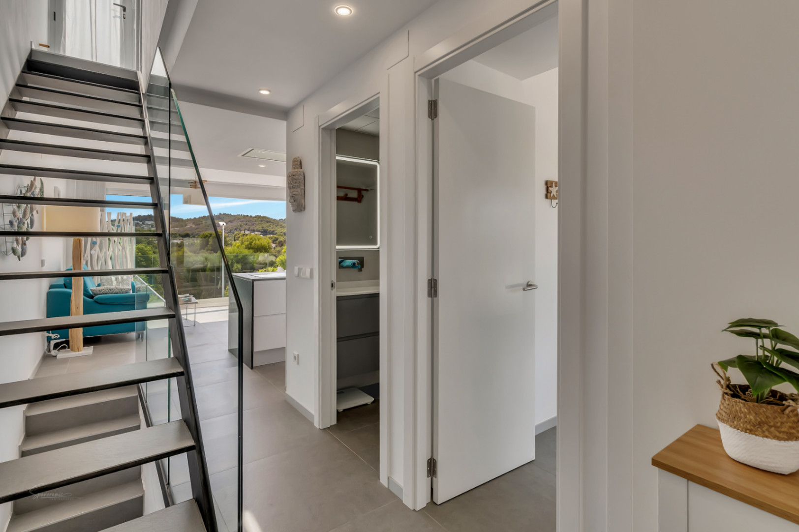 Distribuidor moderno con escalera de peldaños volados y barandilla de cristal. Se aprecia el acceso al baño con espejo retroiluminado y vistas al salón luminoso.