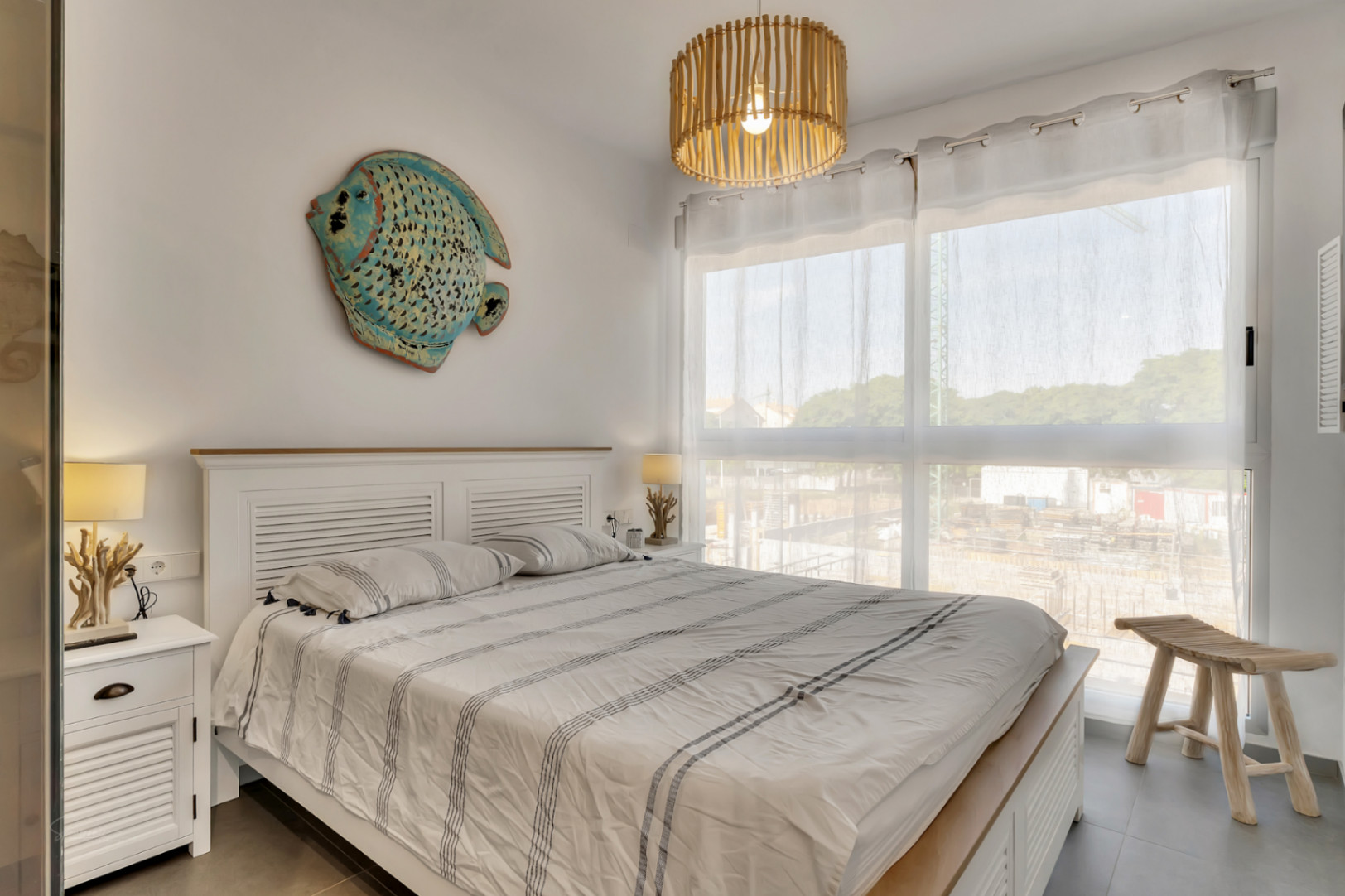 Dormitorio luminoso con cama doble, cabecero de madera blanca y decoración de inspiración marina. Incluye un gran ventanal con luz natural y suelos de gres gris.