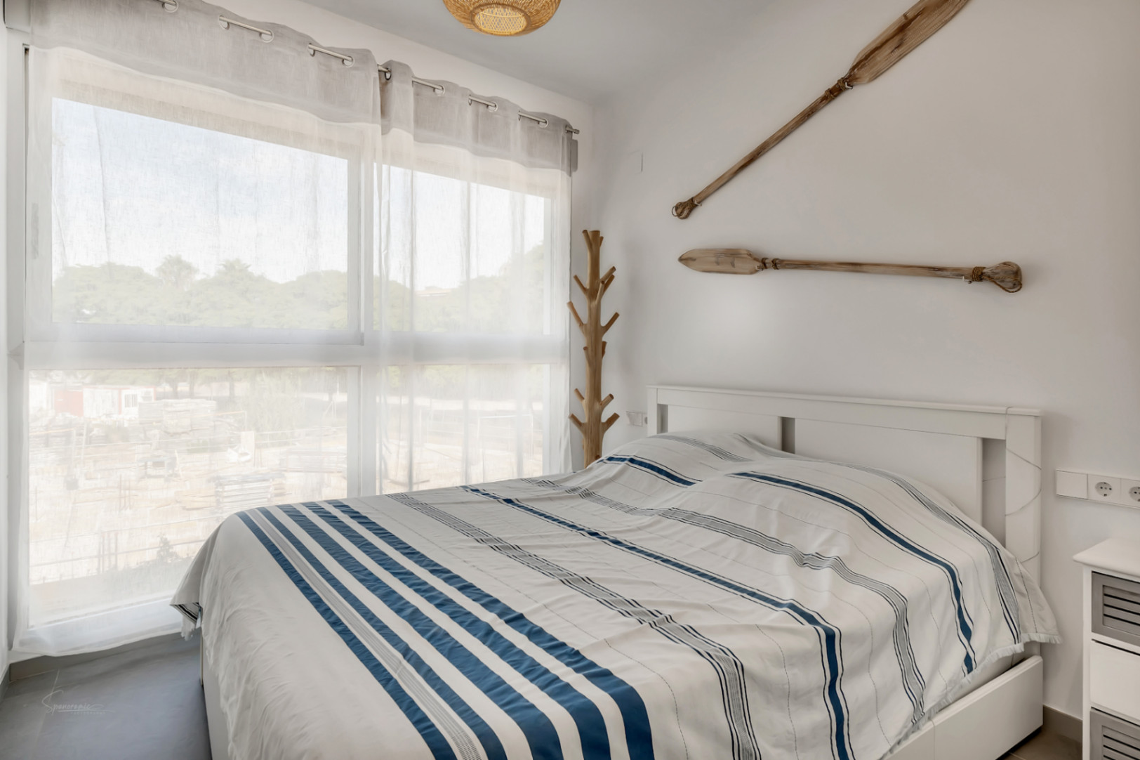 Dormitorio luminoso con cama doble, decoración de inspiración náutica con remos de madera y un perchero artesanal junto al gran ventanal.