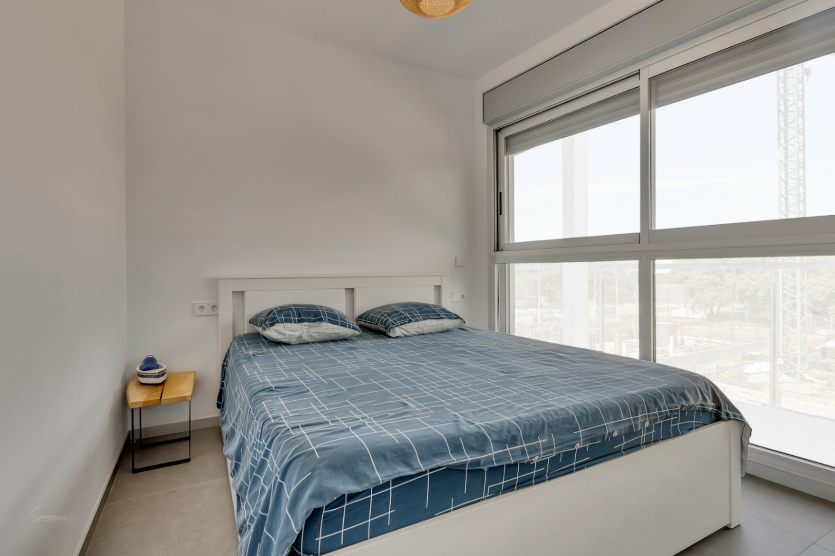 Dormitorio luminoso con cama doble, paredes blancas y gran ventanal de aluminio. Incluye persianas motorizadas y suelos de gres porcelánico en tonos grises.