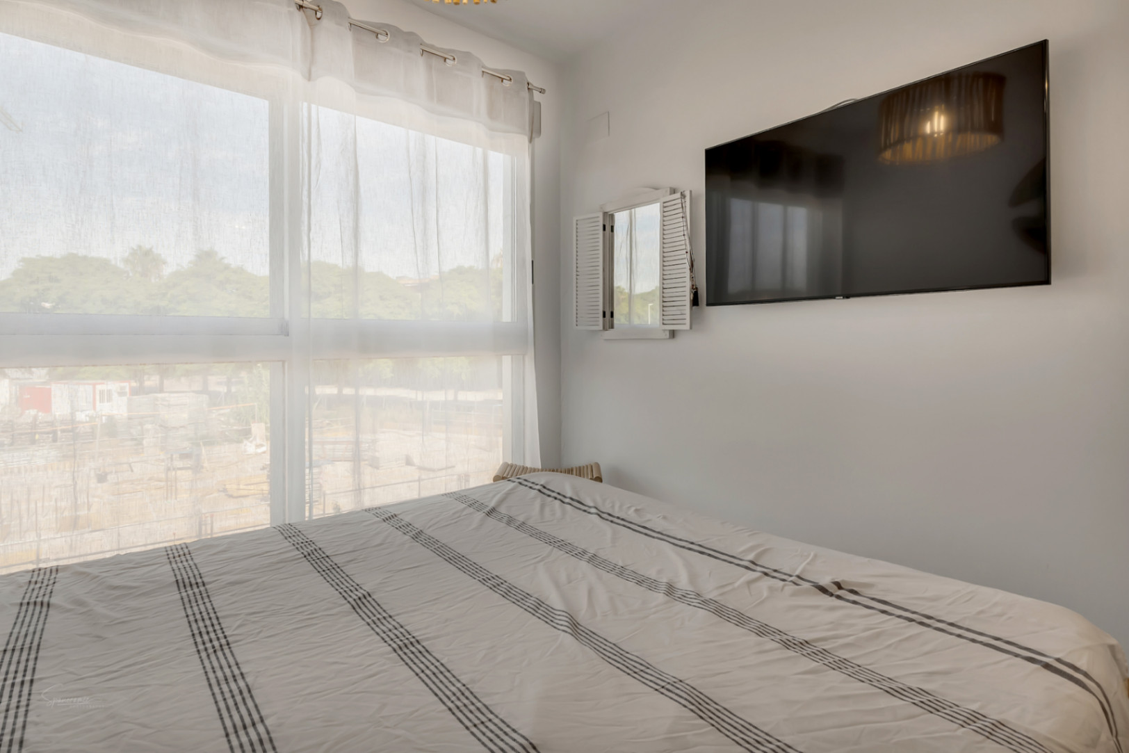 Dormitorio luminoso con gran ventanal, cortinas traslúcidas, cama doble con ropa de cama a rayas y televisión de pantalla plana montada en la pared.