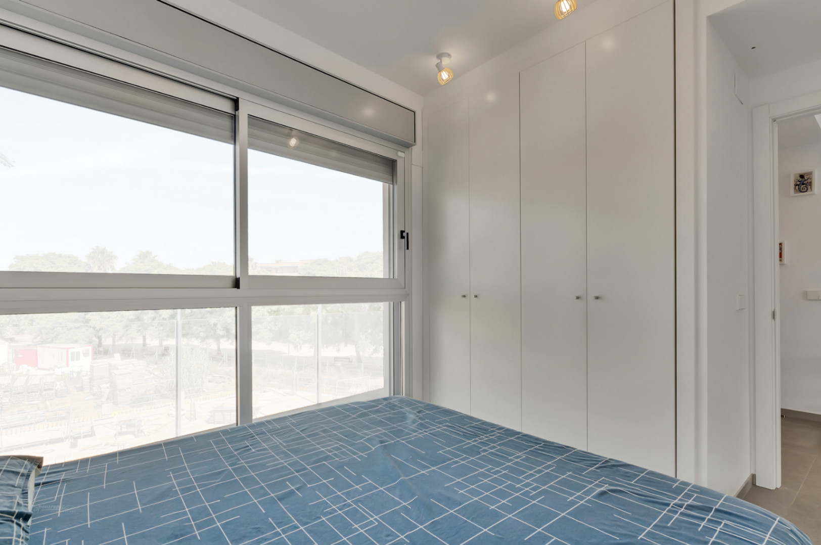 Dormitorio moderno con armarios empotrados blancos de suelo a techo y gran ventanal con persianas motorizadas que ofrece abundante luz natural.