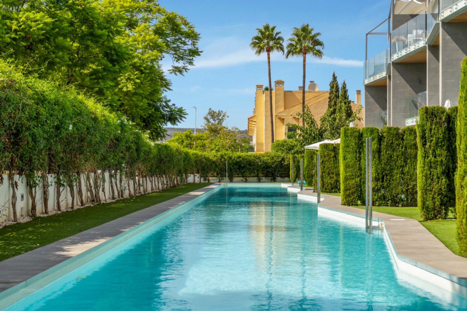 Piscina comunitaria de 53 metros de largo rodeada de vegetación mediterránea, setos cuidados y edificios de arquitectura moderna bajo un cielo despejado.