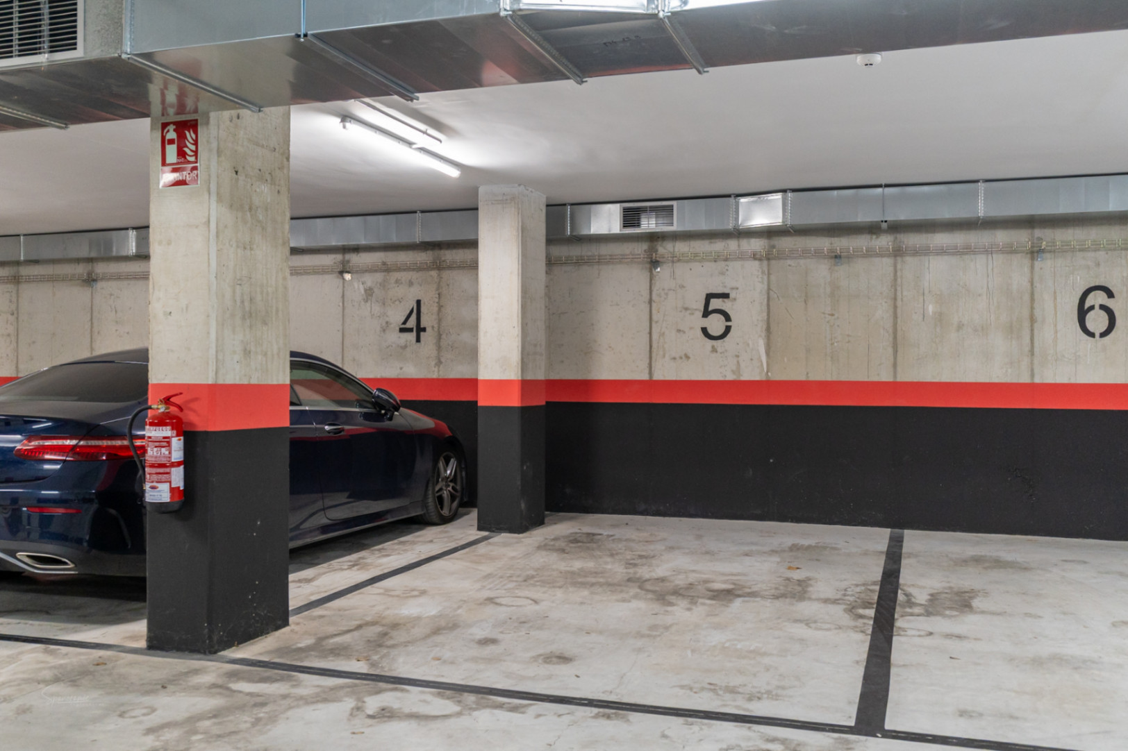 Plaza de garaje subterránea numerada con acabados en hormigón visto, conductos de ventilación industriales y sistemas de seguridad contra incendios integrados.