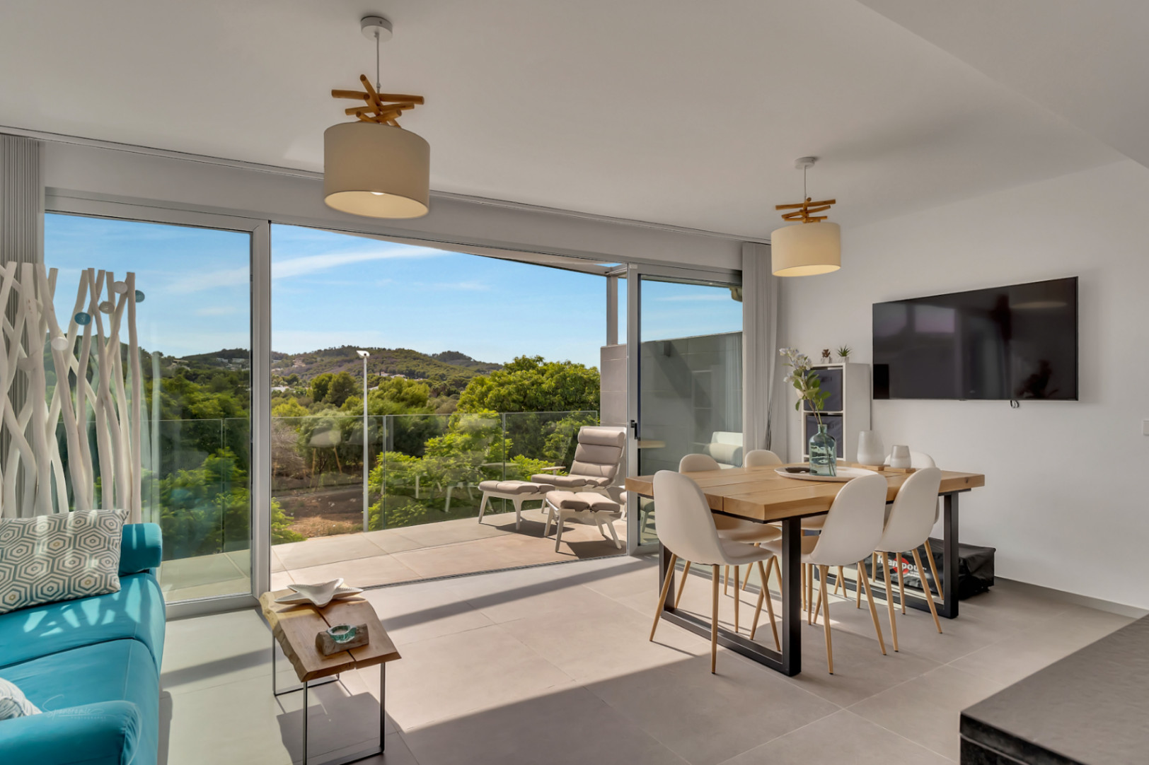 Salón comedor luminoso con grandes ventanales correderos que dan acceso a un balcón con vistas despejadas al campo. Incluye mesa de madera, sillas nórdicas y suelos porcelánicos.