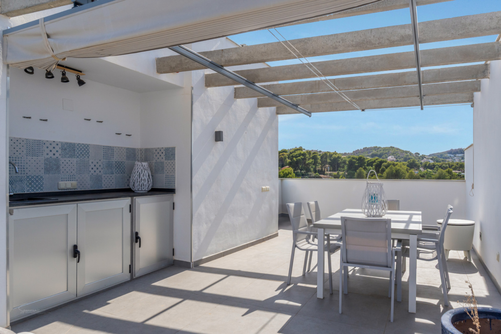 Terraza privada con cocina de verano, encimera oscura y comedor exterior bajo una pérgola de vigas de hormigón con vistas despejadas al entorno natural.