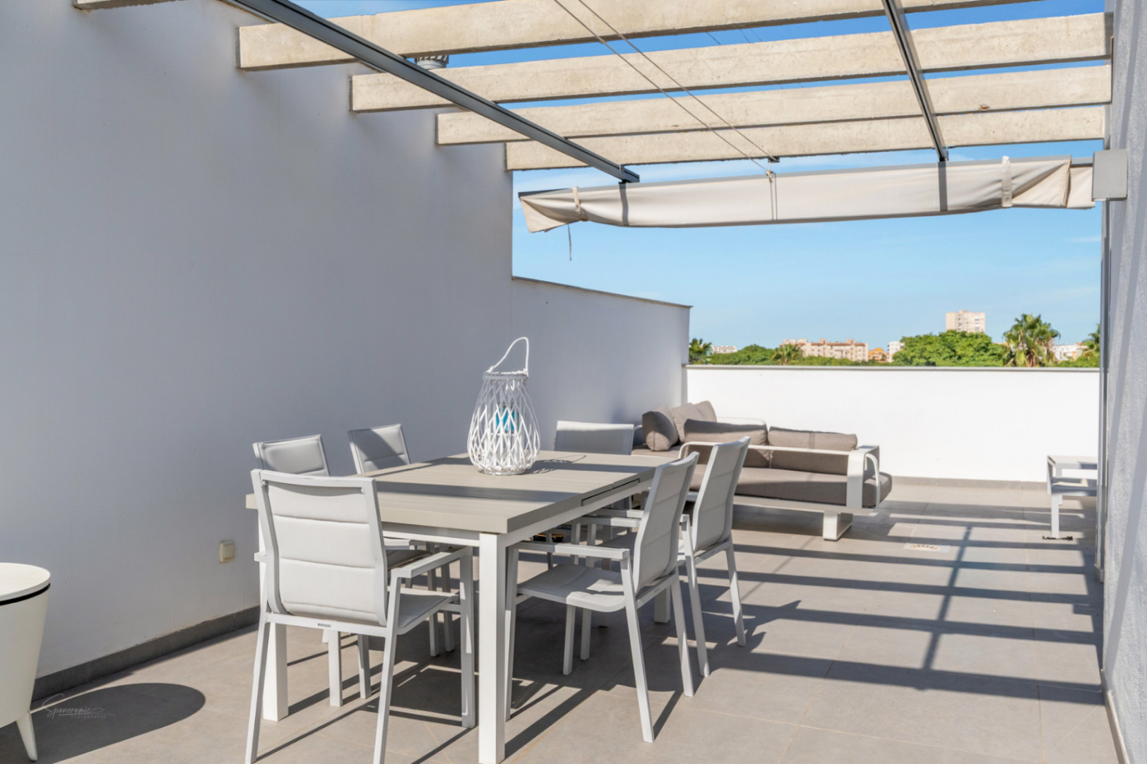 Terraza privada en la azotea con zona de comedor exterior, sofá lounge y pérgola de hormigón con toldo extensible sobre suelo de gres porcelánico.