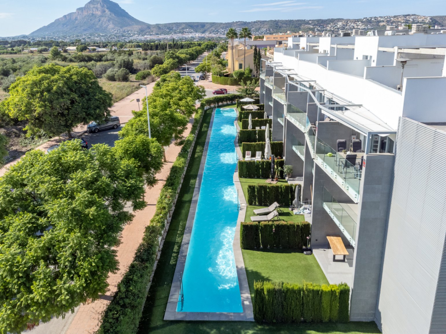 Vista aérea de la piscina comunitaria de 53 metros de largo, rodeada de jardines privados y arquitectura moderna con vistas al Montgó y campos protegidos.