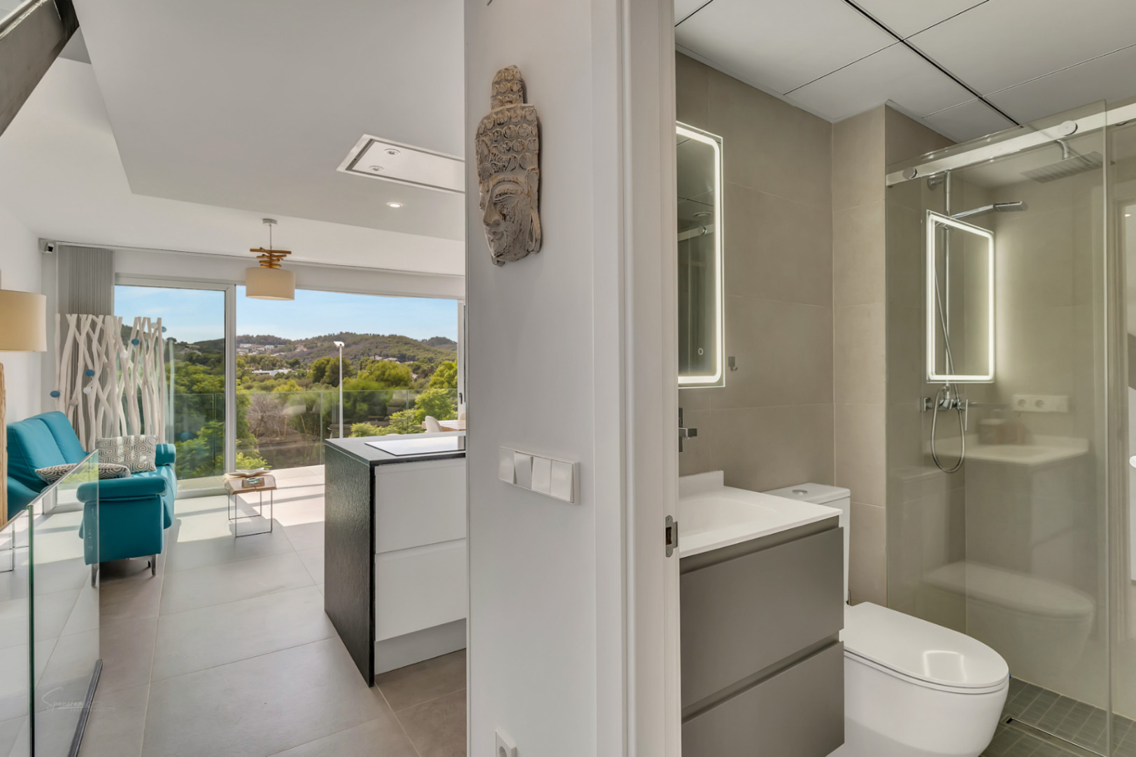 Vista desde el pasillo hacia un baño moderno con espejo retroiluminado y ducha de cristal, conectando con el salón de concepto abierto y vistas exteriores.