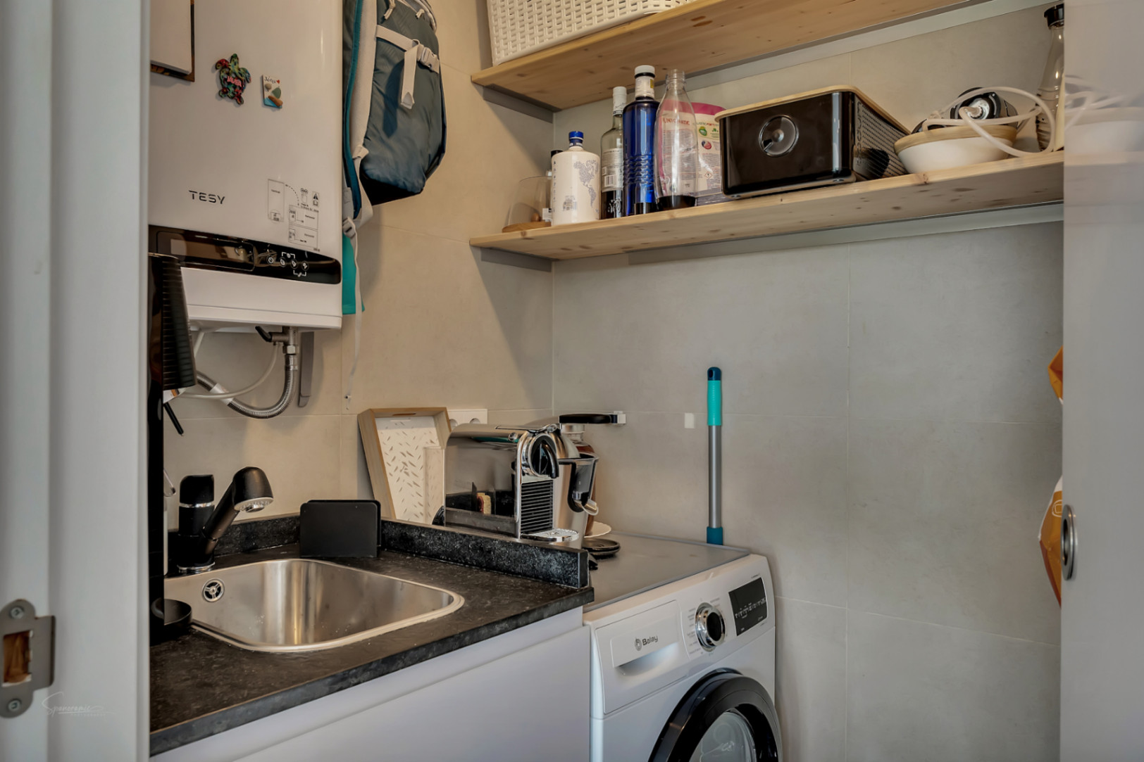 Zona de lavandería funcional equipada con lavadora Balay, fregadero de acero inoxidable, termo eléctrico Tesy y estanterías de madera.