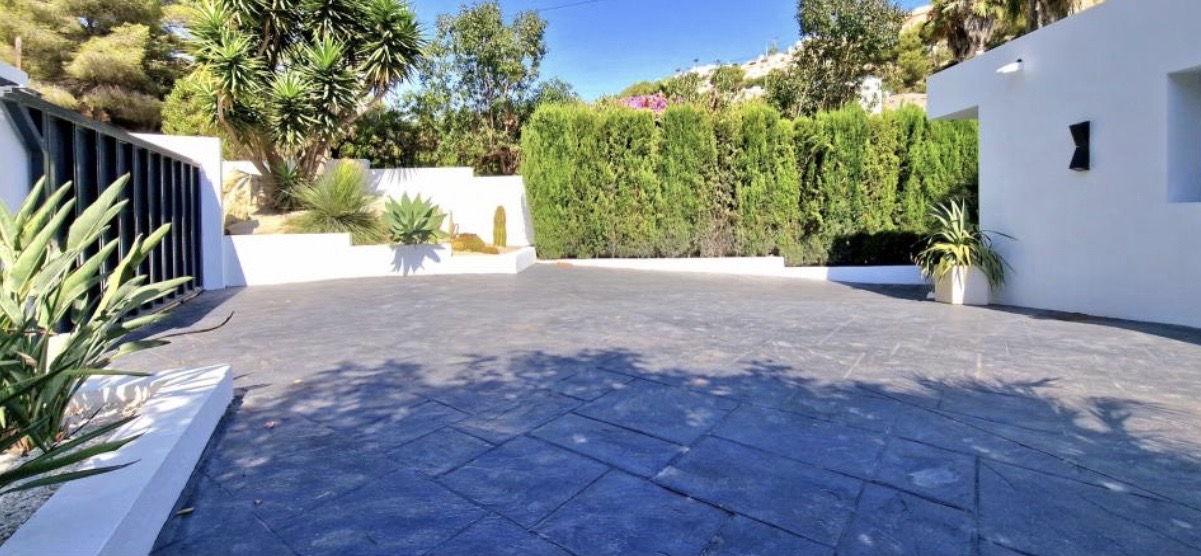 Amplia zona de aparcamiento con pavimento de hormigón impreso oscuro, rodeada de muros blancos y vegetación mediterránea con yucas y setos de ciprés.