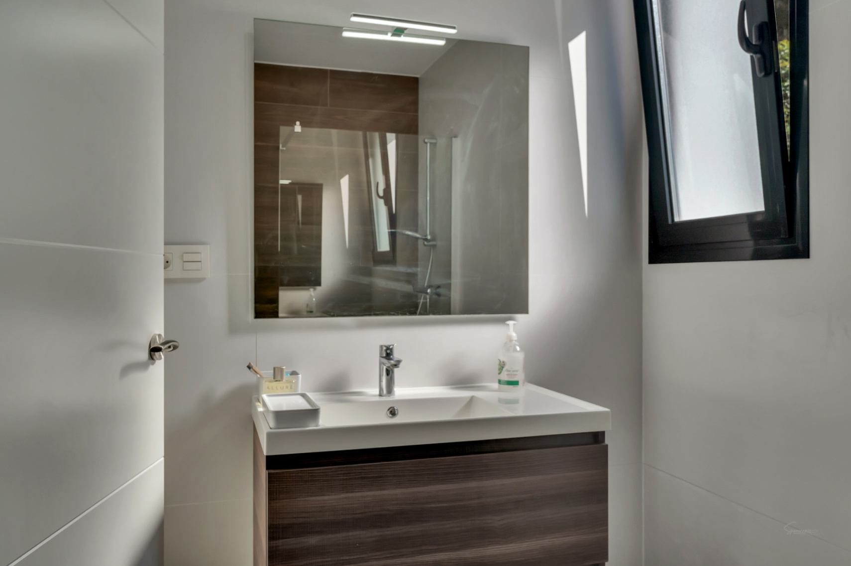 Baño contemporáneo con mueble suspendido en madera oscura, lavabo blanco de líneas rectas y espejo con aplique superior. Incluye ventana de carpintería negra.