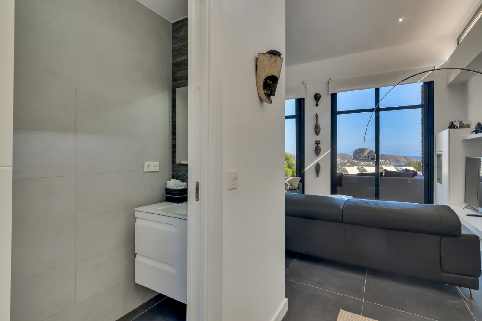 Baño de cortesía con lavabo suspendido junto al salón principal, que destaca por sus amplios ventanales con vistas al mar y decoración contemporánea.