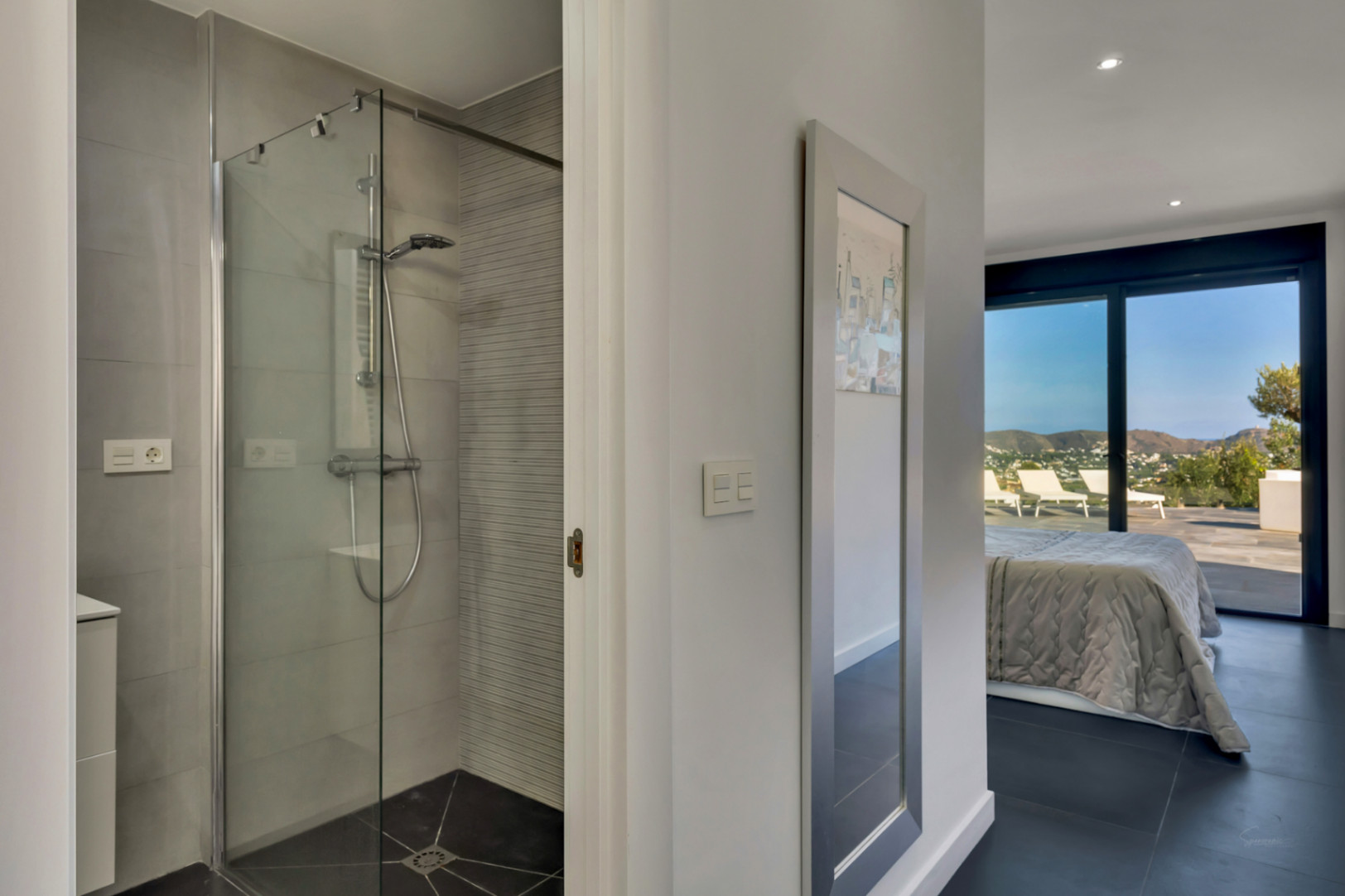 Baño en suite con ducha de obra acristalada y azulejos grises, conectado a un dormitorio con amplios ventanales y vistas despejadas a la montaña.