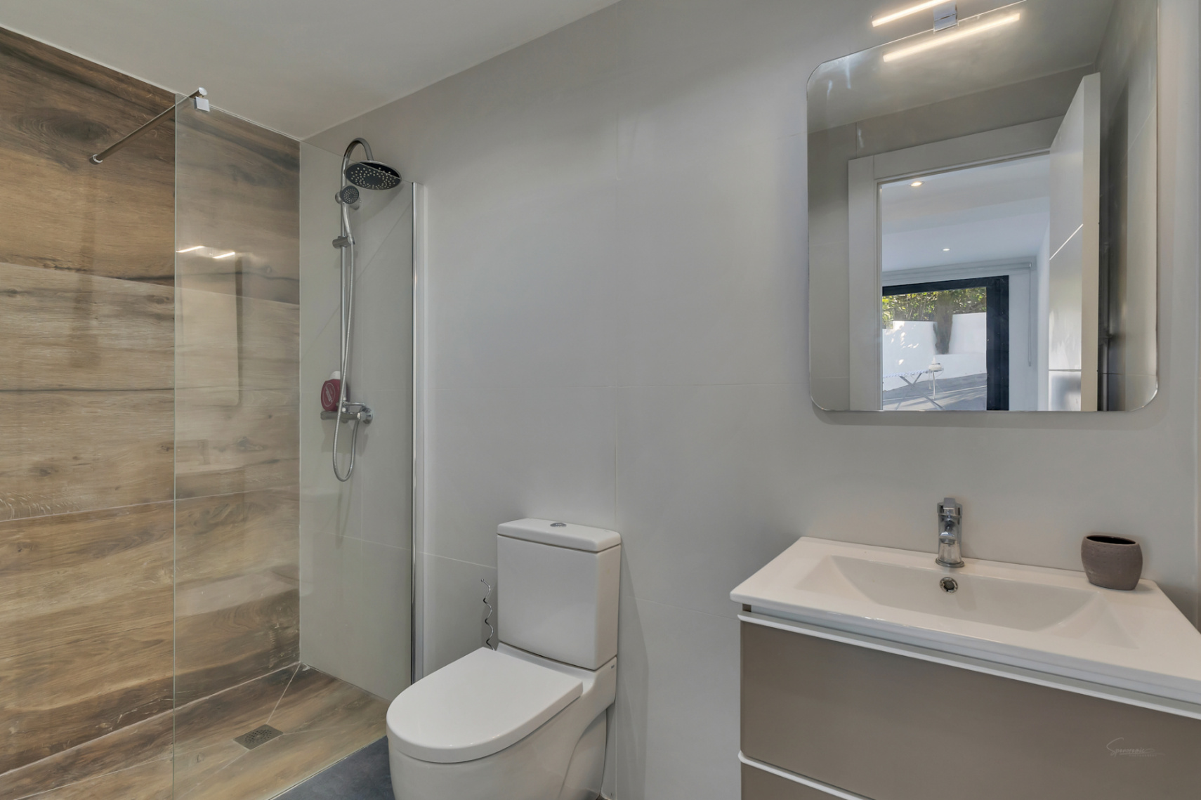 Baño moderno con ducha a ras de suelo revestida en gres porcelánico efecto madera, mampara de cristal fija y mueble de lavabo suspendido con espejo iluminado.