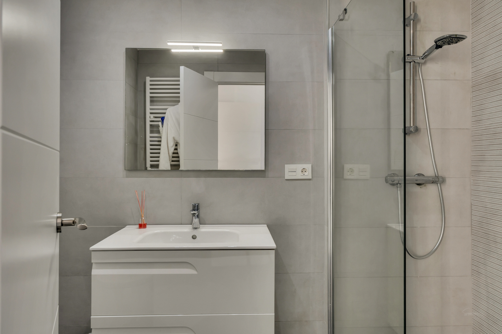 Baño moderno con mueble de lavabo blanco, espejo con iluminación superior y ducha con mampara de cristal y azulejos en tonos grises.